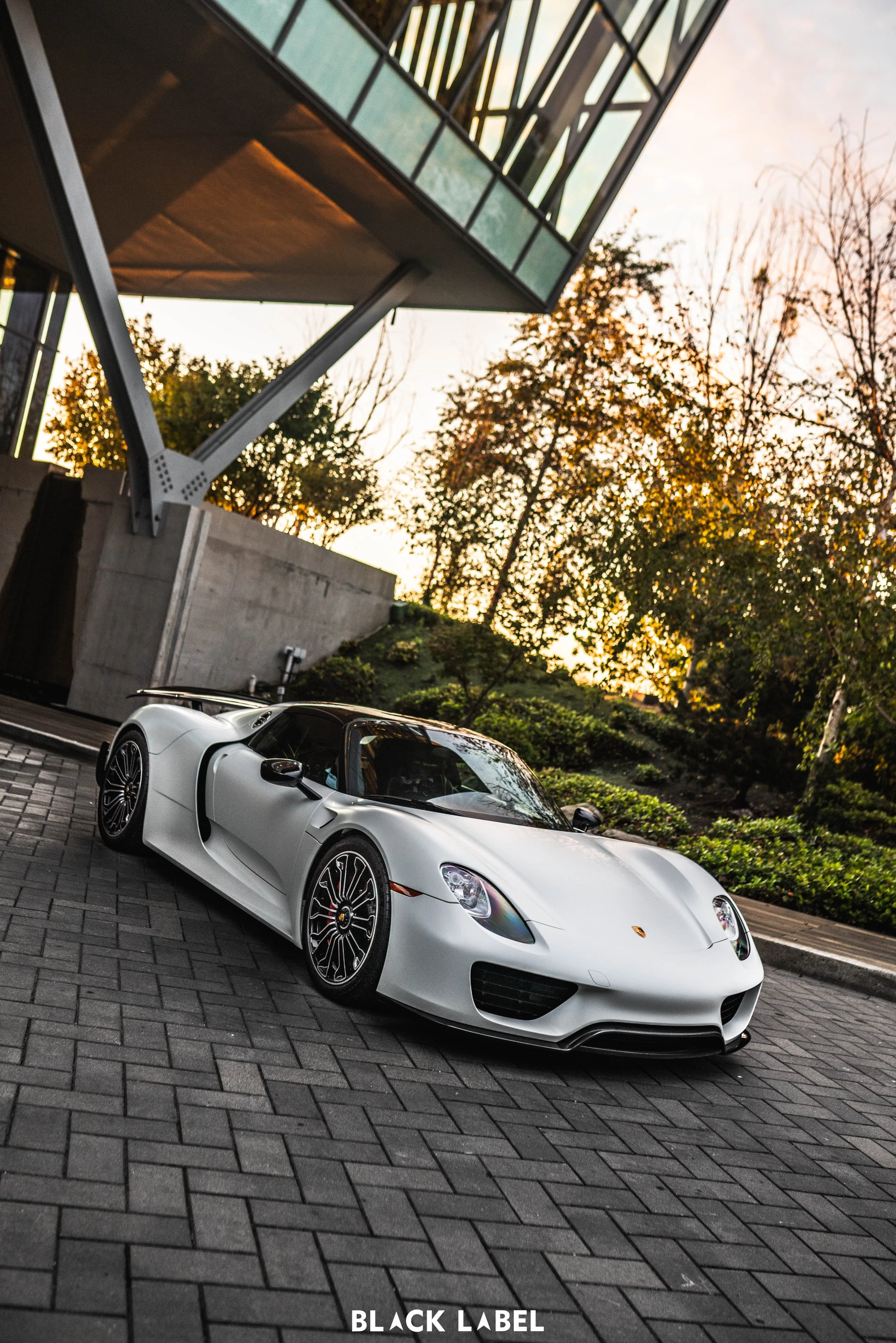 A white Porsche supercar parked on a paved brick driveway near a modern building with glass windows and metallic supports, with trees and shrubs in the background during sunset.