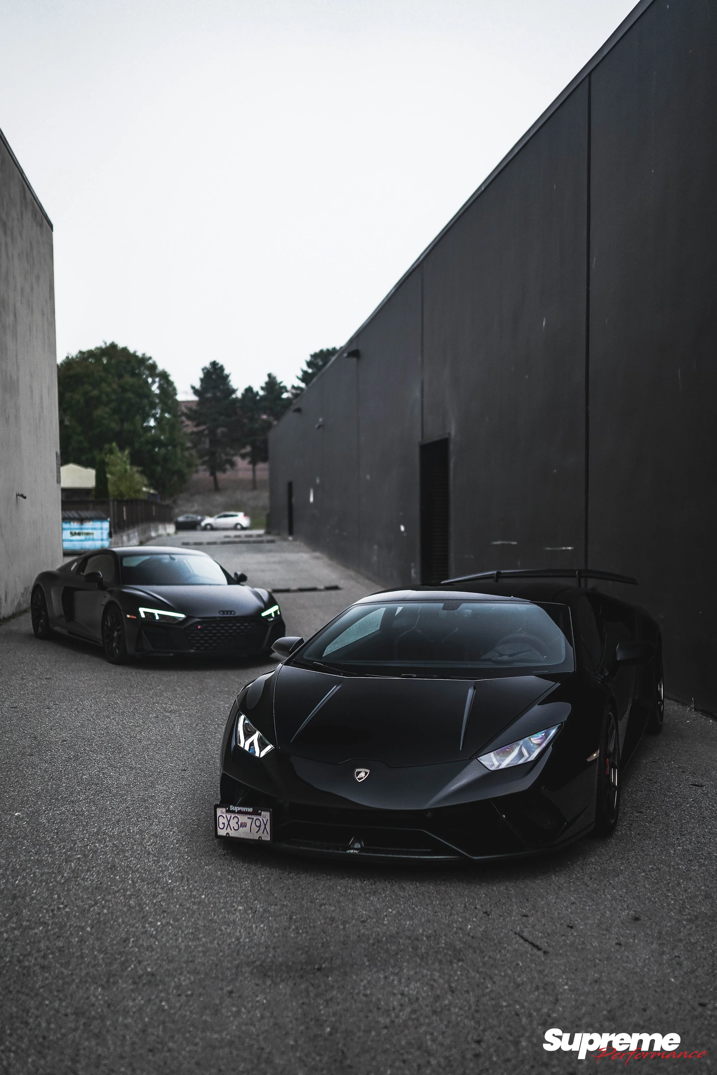 Two black luxury sports cars, a Lamborghini and an Audi R8, parked in an alleyway near black building walls with trees in the background. A 'Supreme Performance' logo is in the bottom right corner.