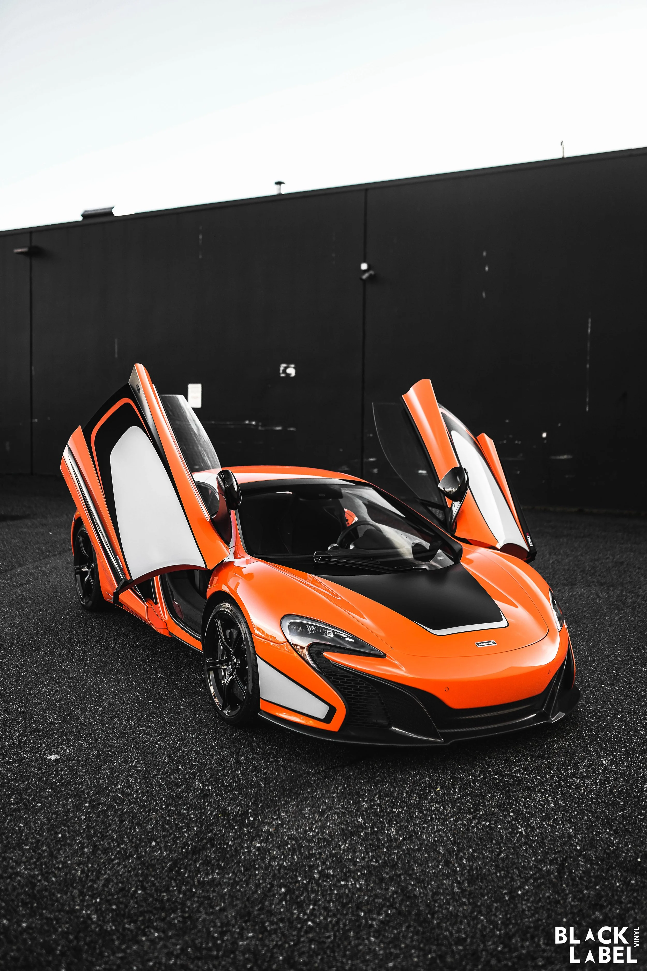 An orange sports car with distinctive black and white accents, with its gullwing doors open, parked on a black textured surface against a dark wall.