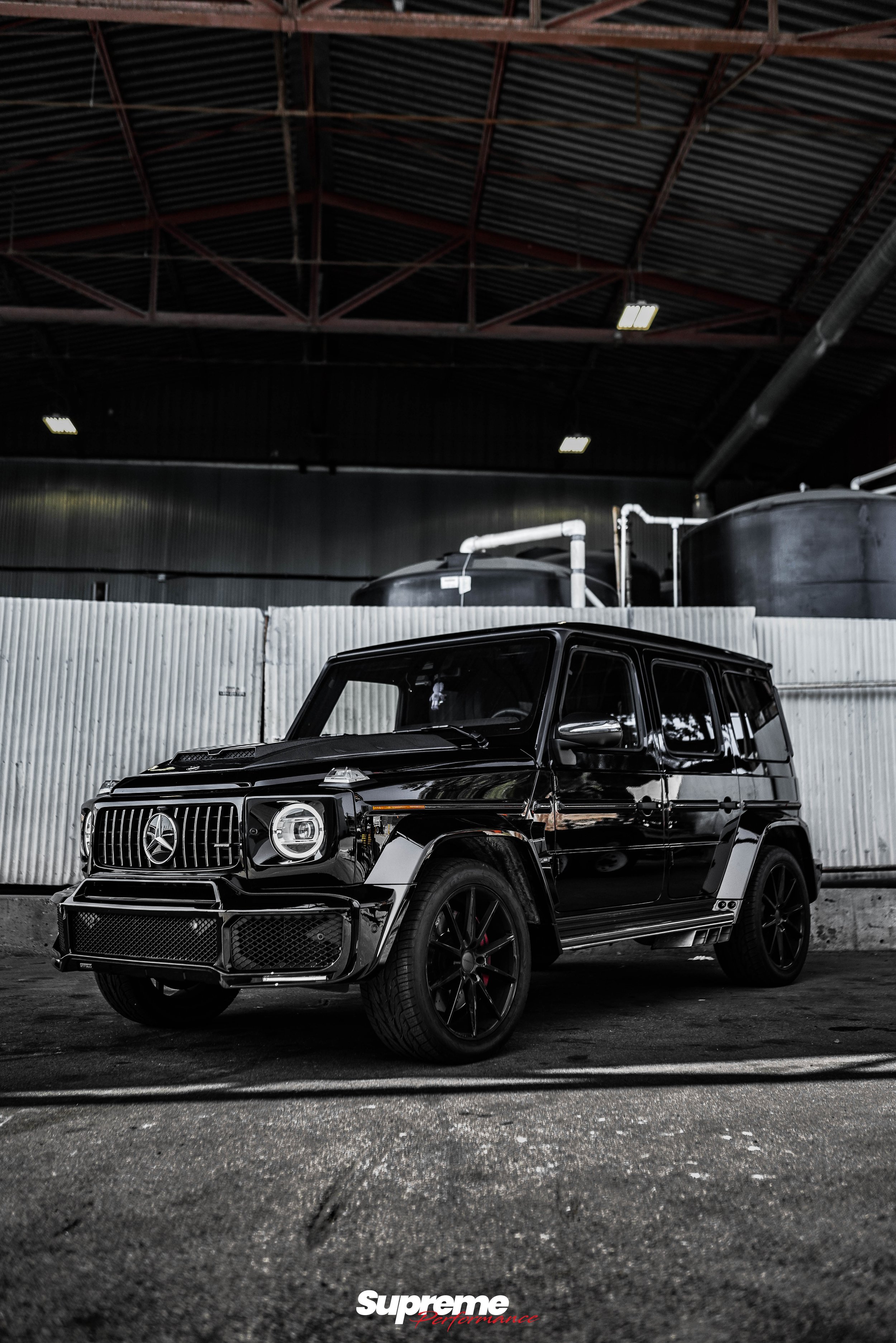 A black Mercedes-Benz G-Class SUV parked inside an industrial or warehouse space with a corrugated metal wall in the background and large black tanks above.
