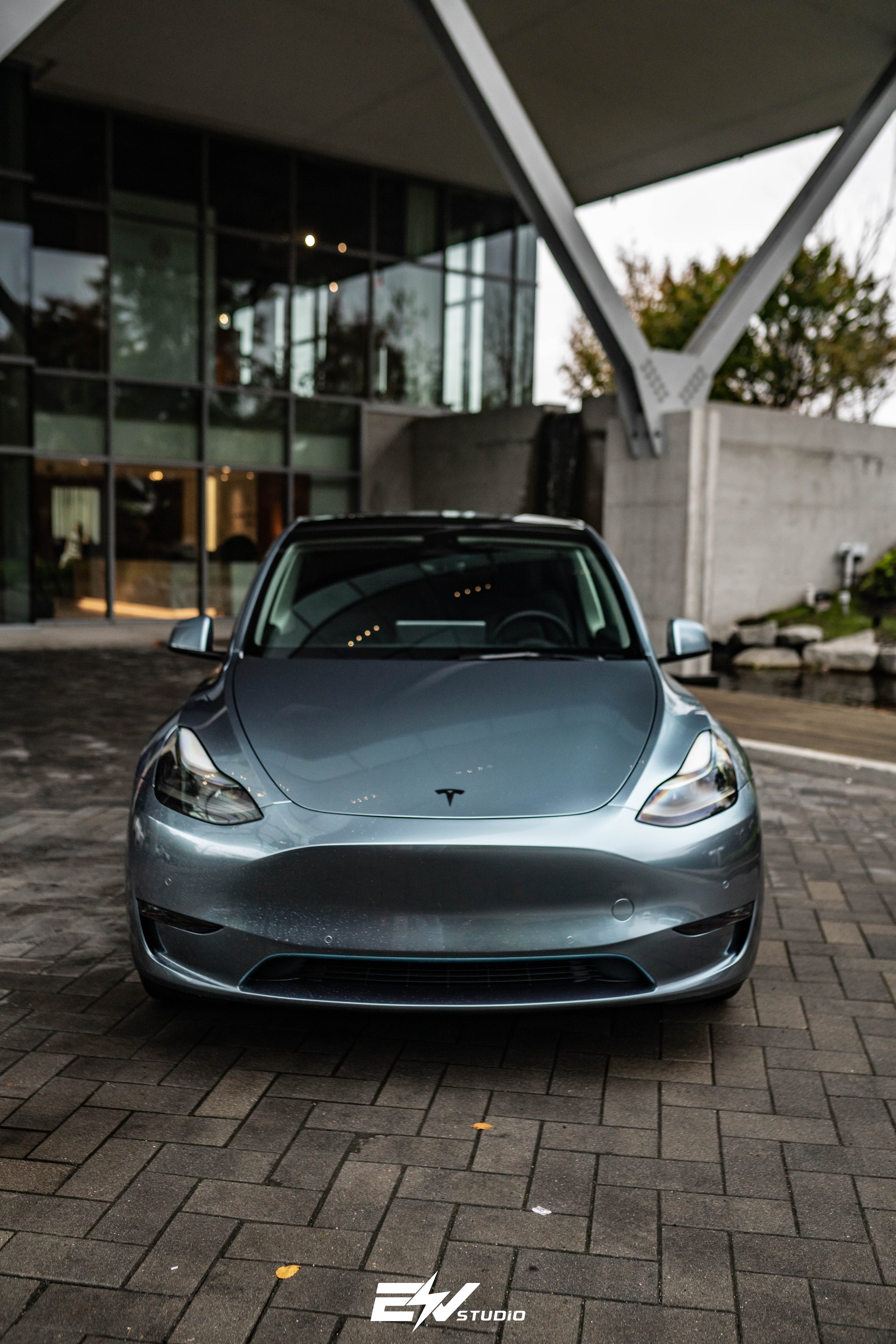 Front view of a silver Tesla Model 3 parked on a brick driveway outside a modern building with large glass windows and trees in the background.