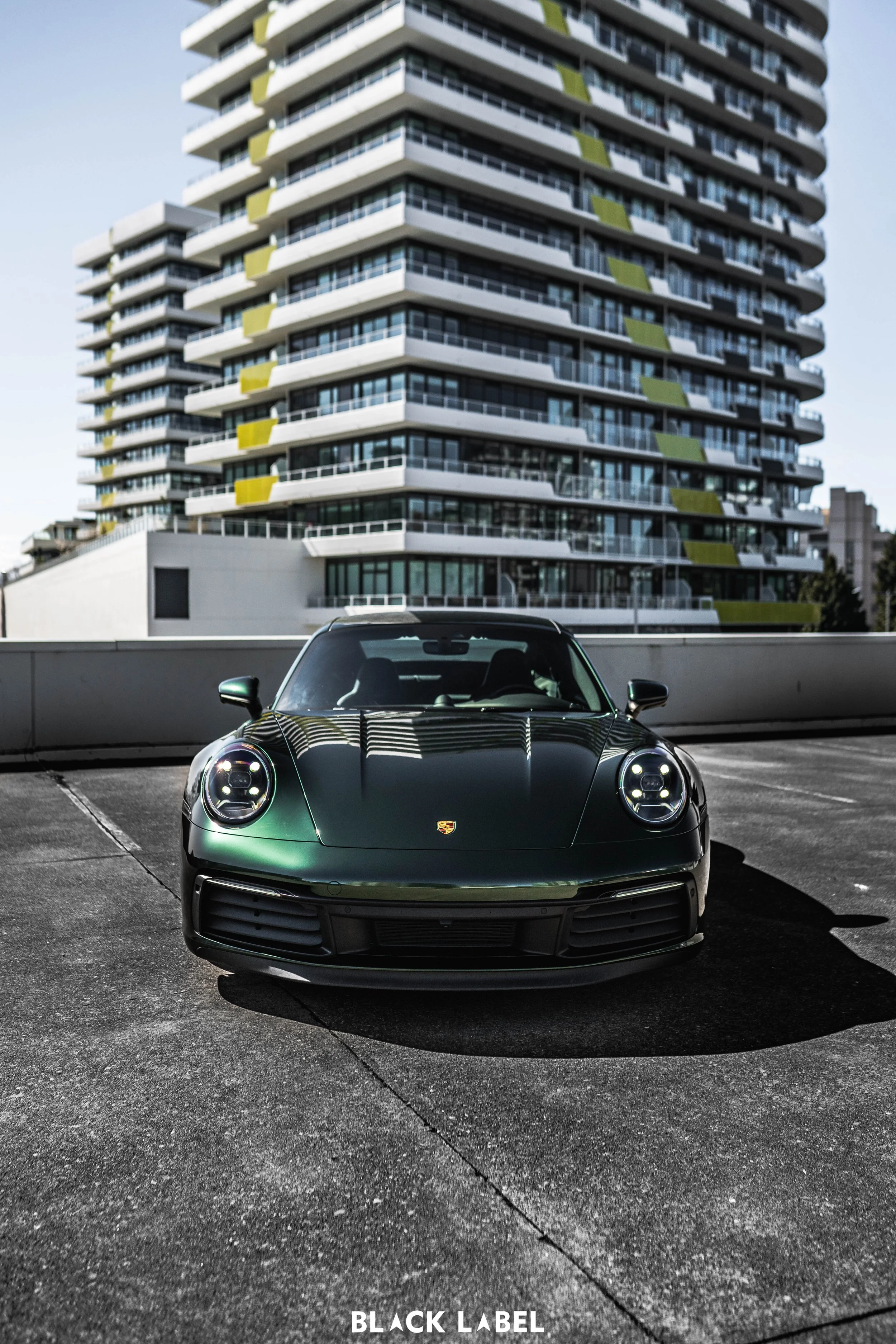 A black Porsche sports car parked on a rooftop parking lot with a modern apartment building in the background.