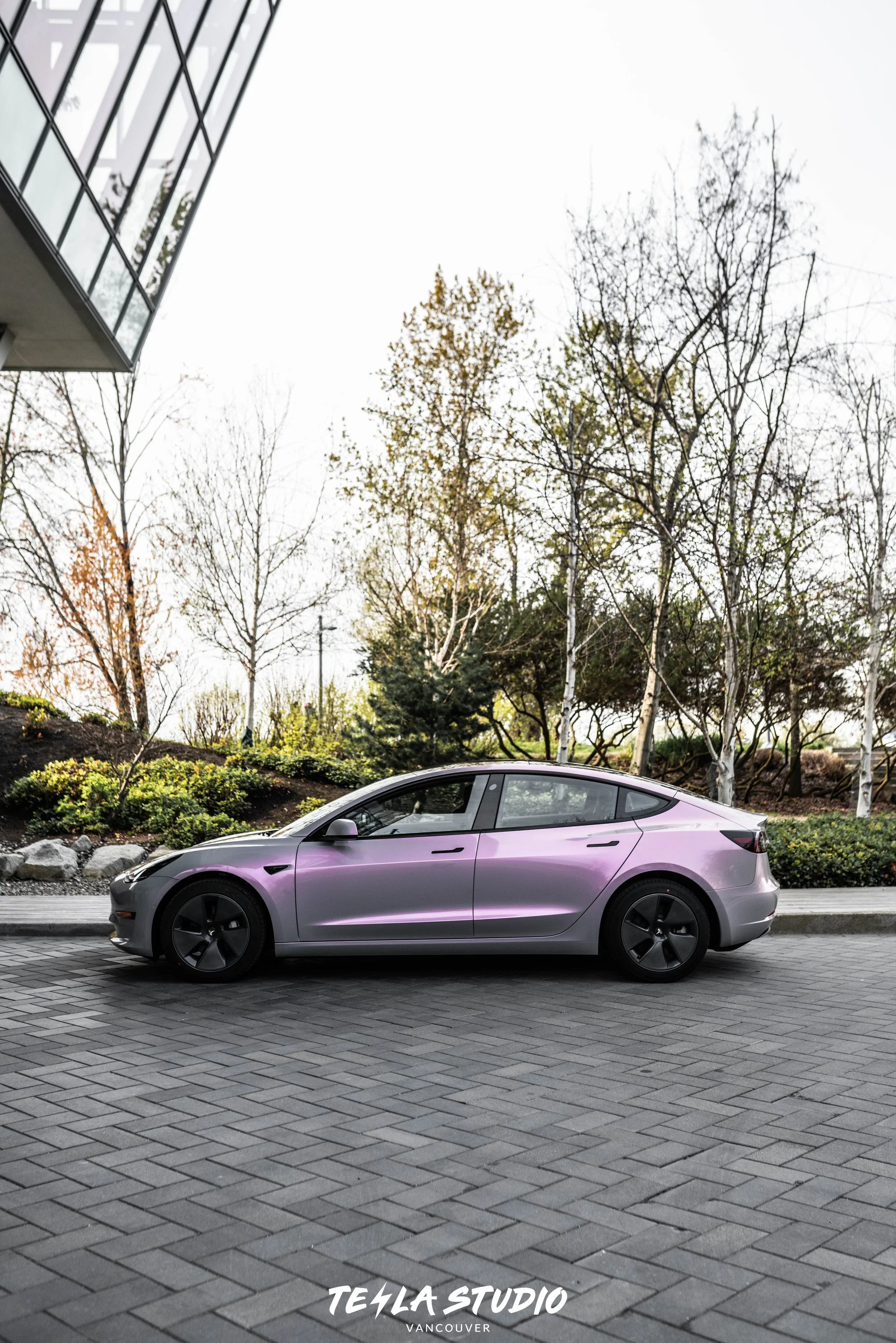 A purple Tesla Model 3 parked on a brick pavement with trees and a building with reflective glass windows in the background.