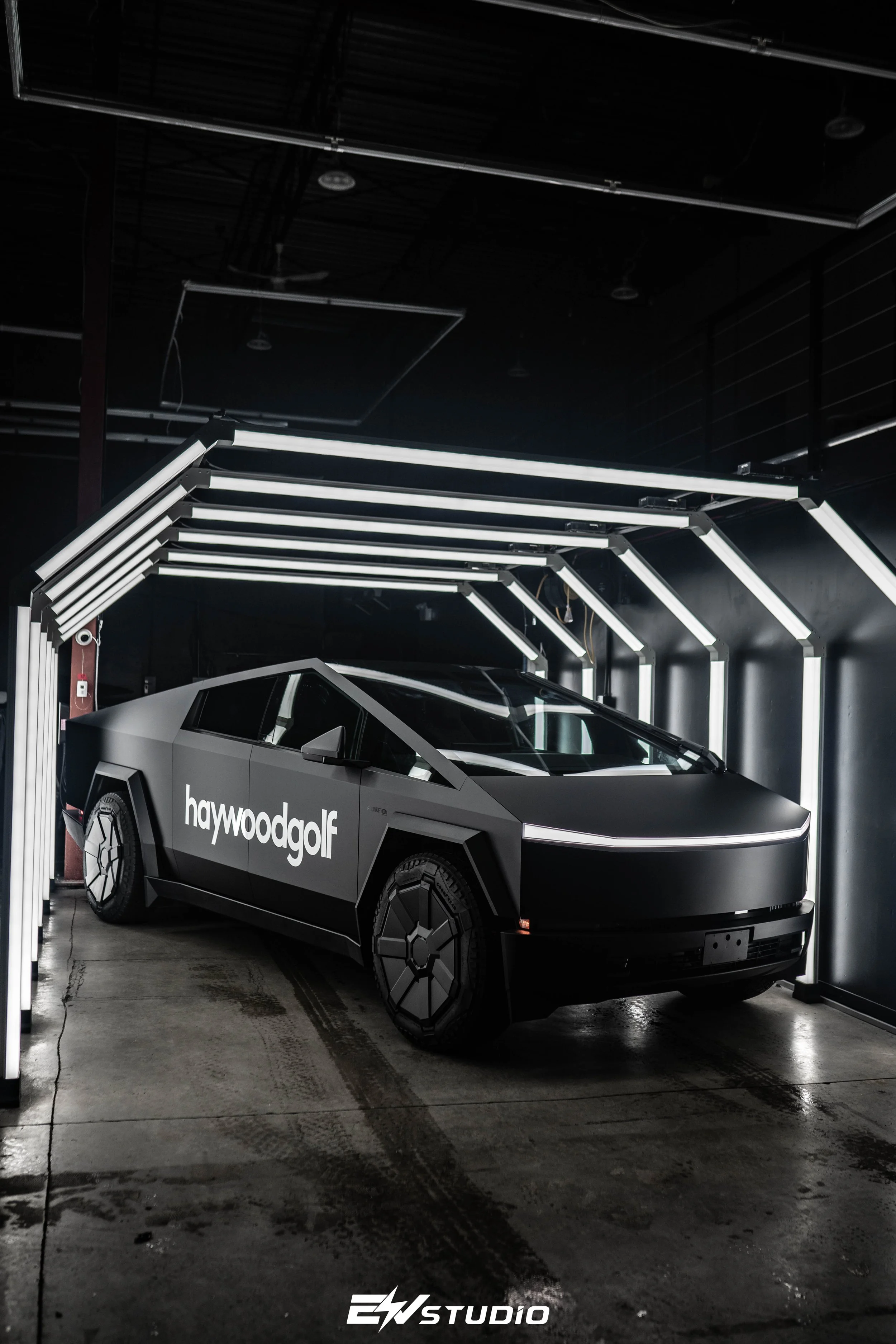 A futuristic black car with the logo 'haywoodgolf' on its side, parked inside a modern, dark showroom with illuminated white geometric lighting structures.