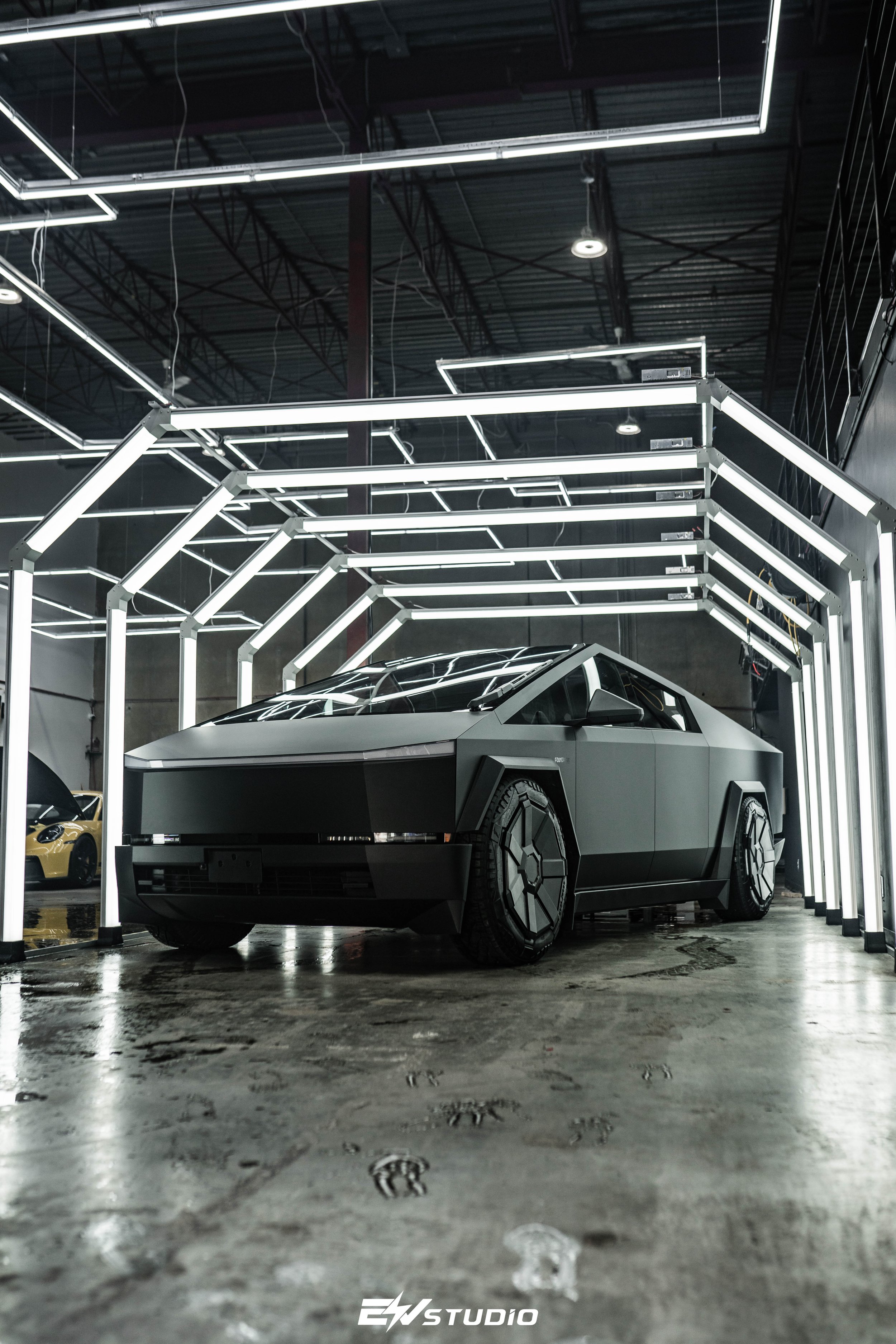 A futuristic, matte gray car with angular design and hexagonal wheel patterns inside a modern, dark showroom with illuminated geometric frame structures overhead.