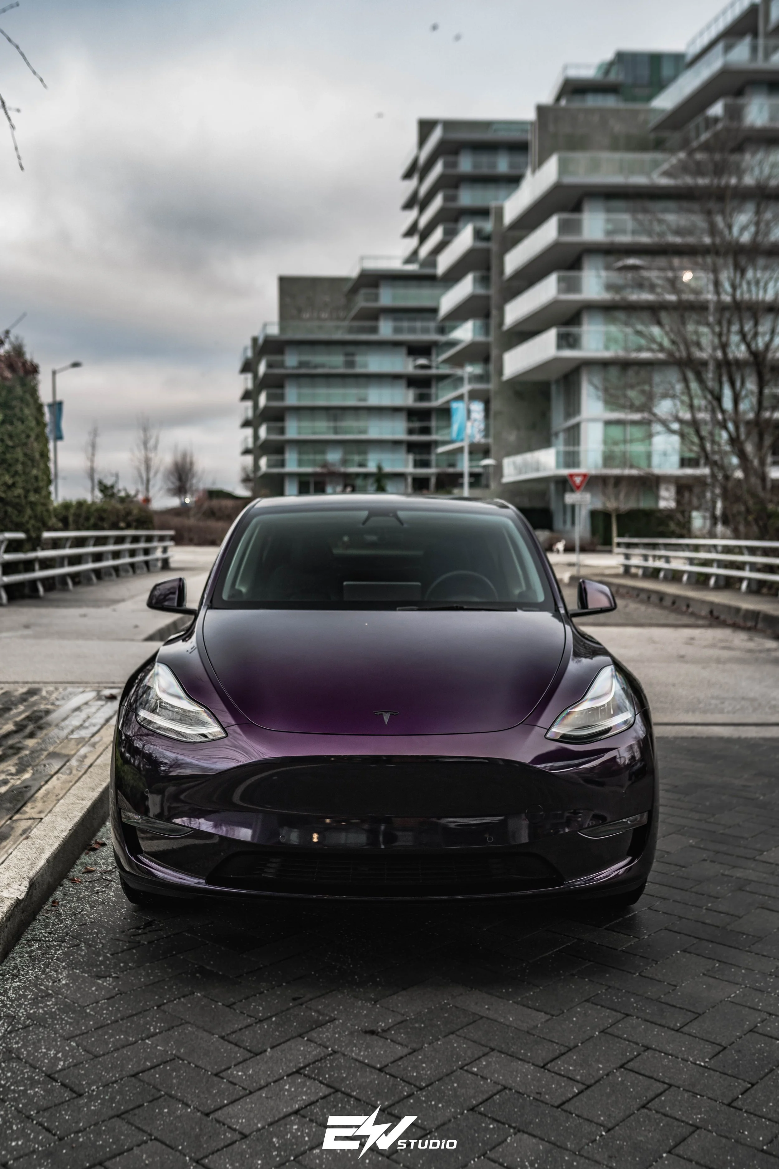 Black Tesla Model 3 parked on a brick street in front of a modern apartment building under a cloudy sky.