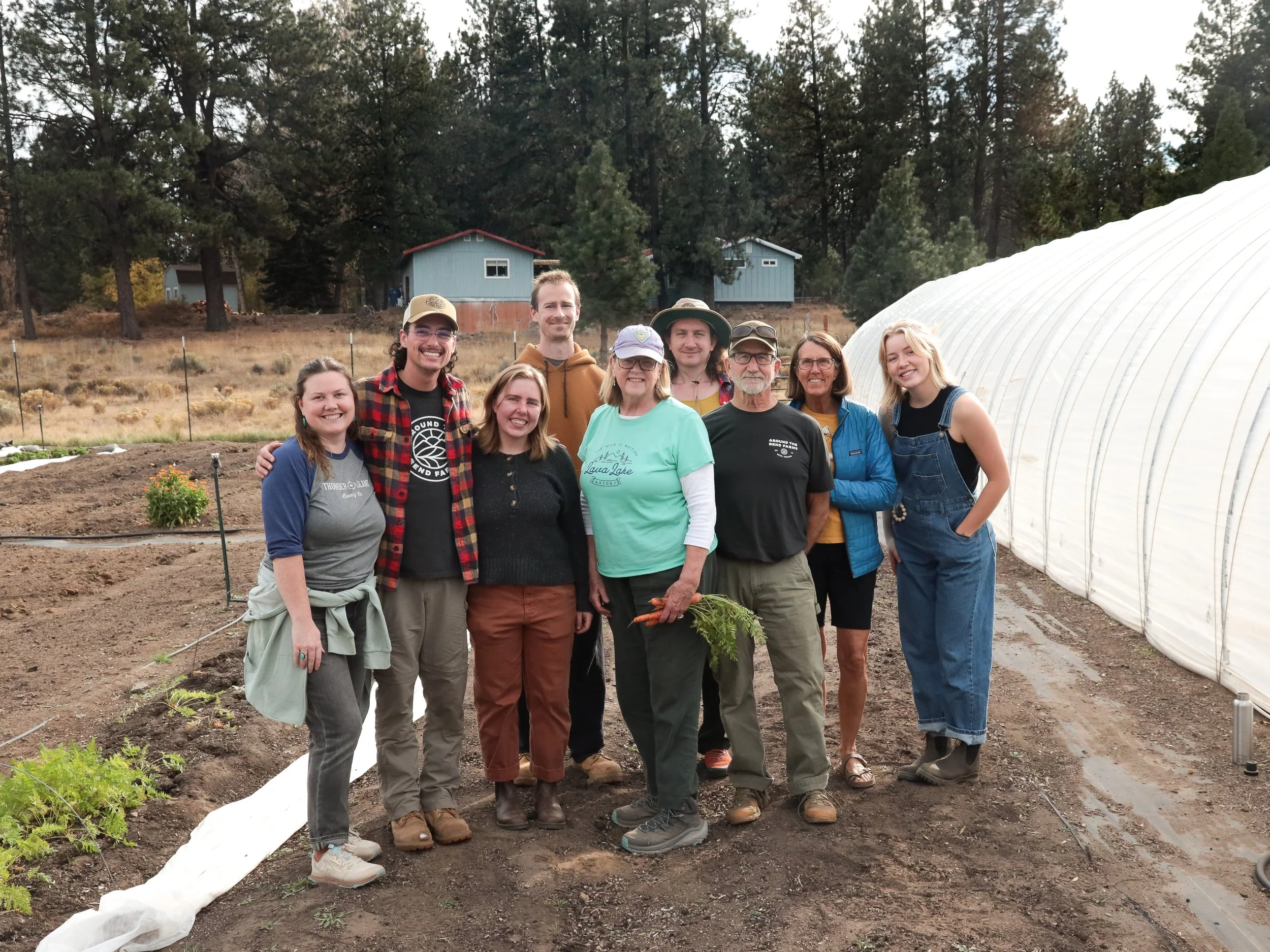 A group of eight people standing together on a farm, with greenhouses and trees in the background.