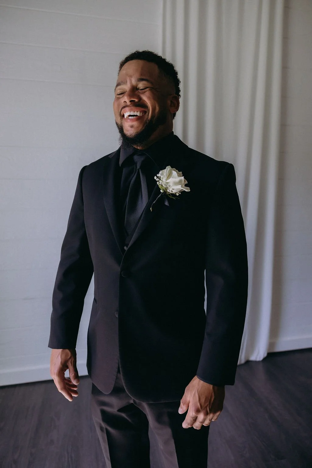 08-groom-laughing-in-a-black-tuxedo-and-black-shirt-with-a-white-rose.jpg