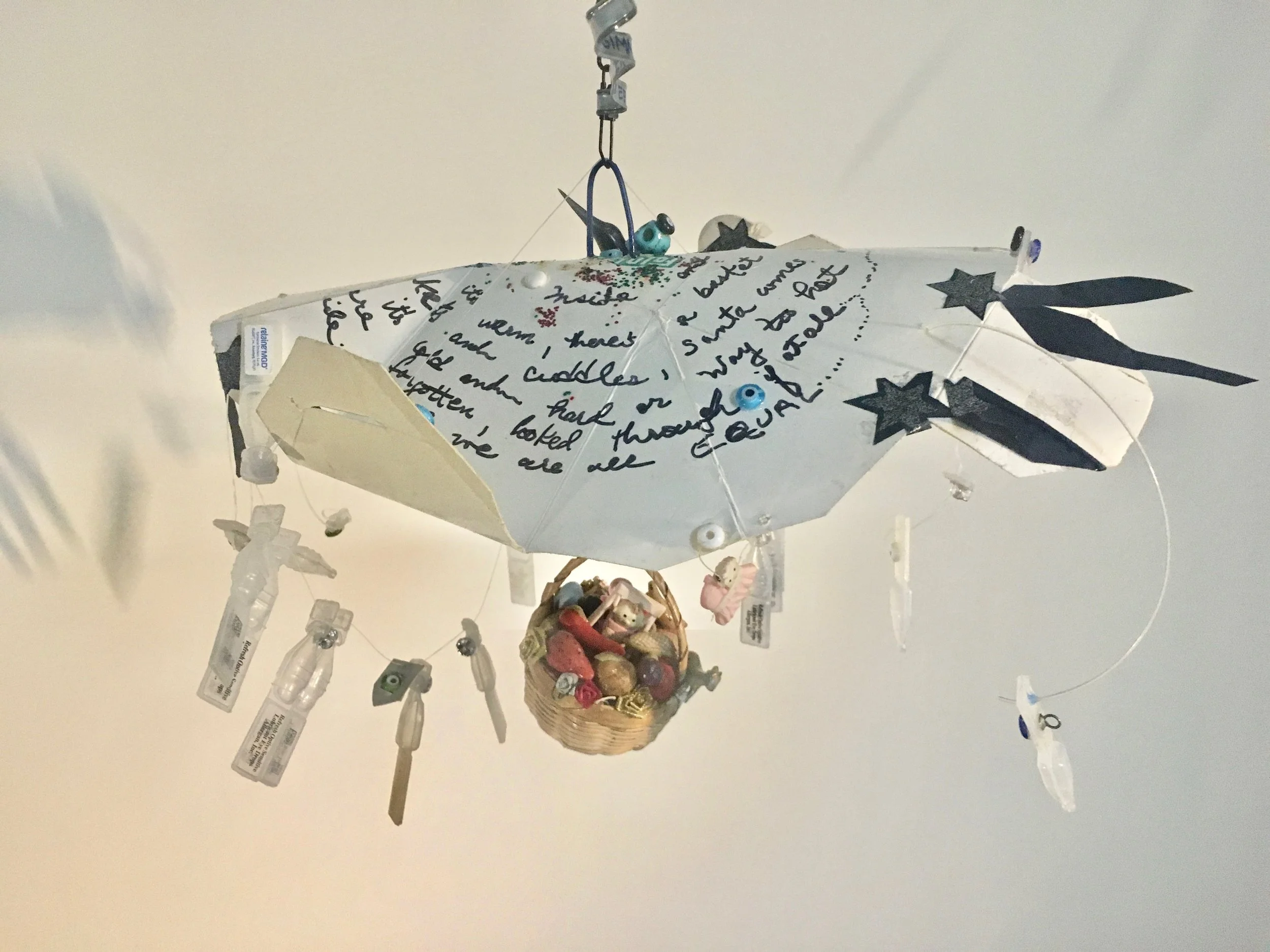 Mixed Media Mobile, “Gimmee Shelter” 10 ‘ long x 11”wide Above the tent is written the following text in a spiral: Inside it’s warm; there’s a basket of plenty of food. There are blankets and cuddles and Santa comes regularly. It’s home, Home, HOME. Outside it’s cold or way too hot and scary, dirty. We are forgotten, looked through, if at all. Death comes wherever we are. Inside and Outside WE are EQUAL.