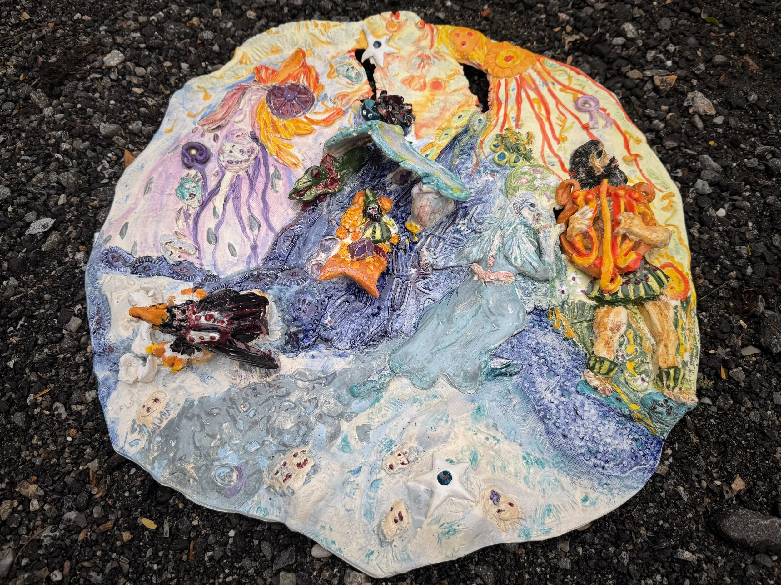 An artistic clay or ceramic mosaic of a mythological or fantasy scene with colorful figures, animals, and creatures, placed on dark gravel ground.