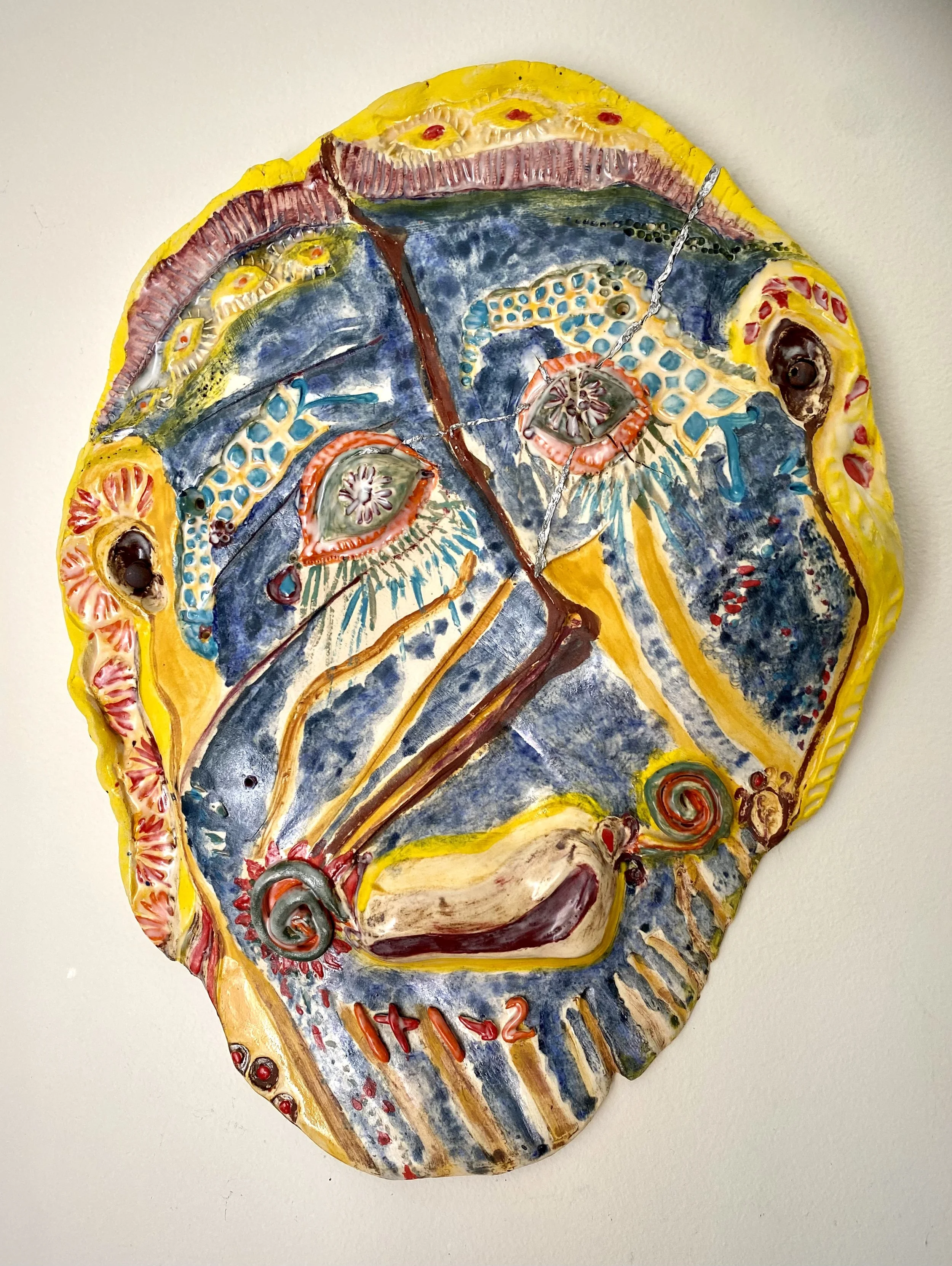 Colorful abstract ceramic mask with eye shapes, spirals, and vibrant patterns in yellow, blue, red, and black.