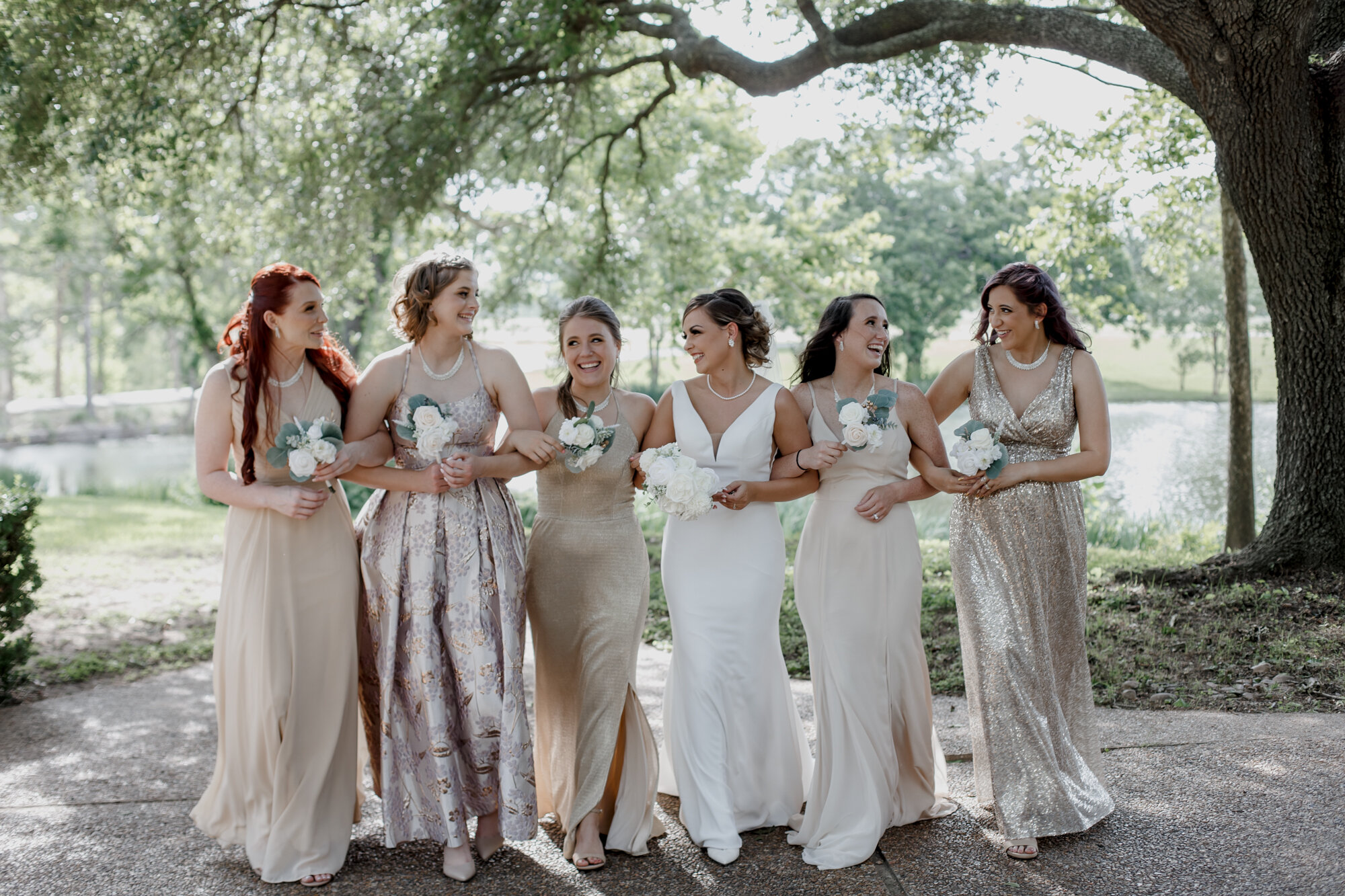 Bride and bridesmaids walking looking at each other. Glowing Wedding in Golden Champagne Tones at The Orchard at Caney Creek in Wharton, TX