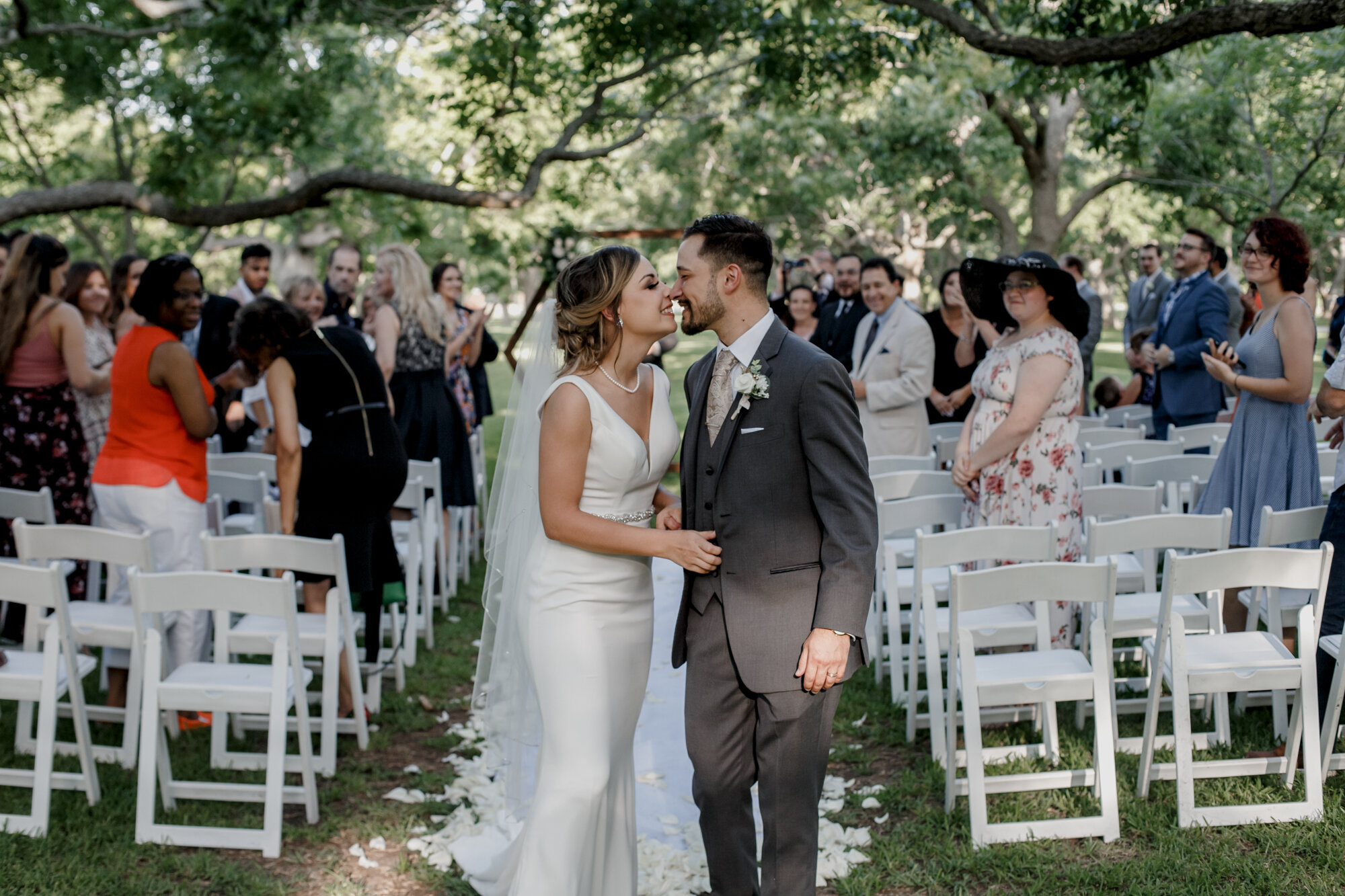 Bride and groom hug and kiss on the aisle. Glowing Wedding in Golden Champagne Tones at The Orchard at Caney Creek in Wharton, TX