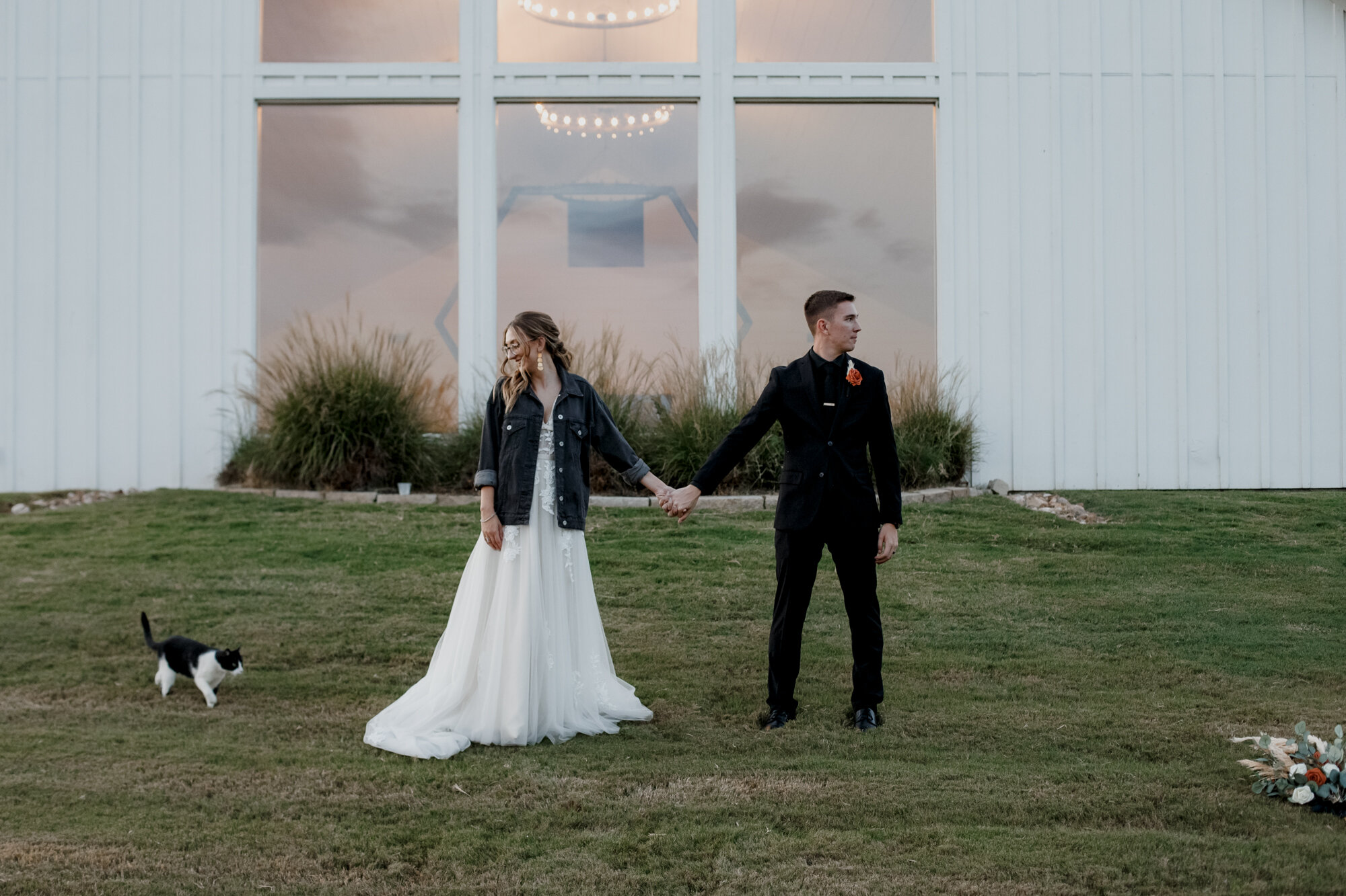 Bride and groom hold hands looking away from each other with a cat. Burnt Orange and Black Fall Wedding at The Farmhouse