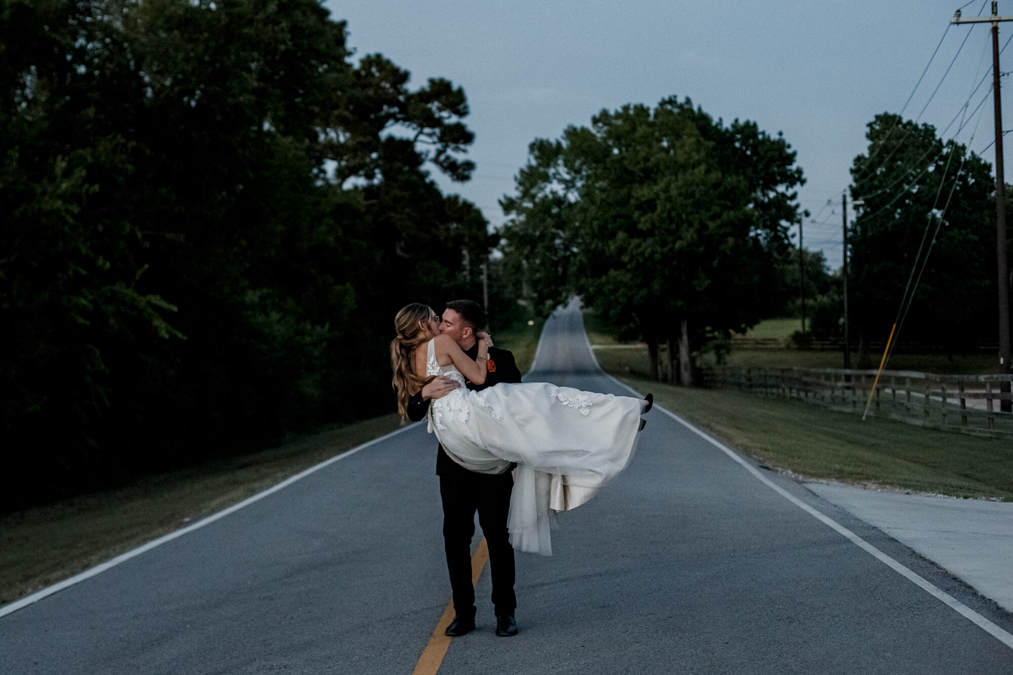 Groom carrying bride walking on the country road. Burnt Orange and Black Fall Wedding at The Farmhouse