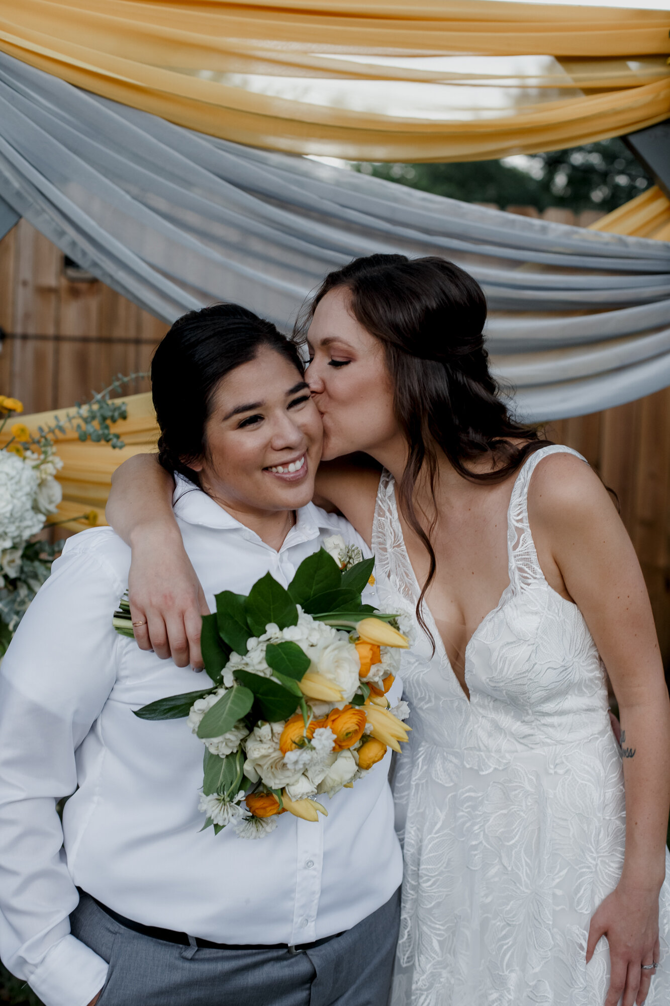 Cozy Laid-Back LGBTQ+ Backyard Wedding. Bride kisses bride holding yellow flowers.