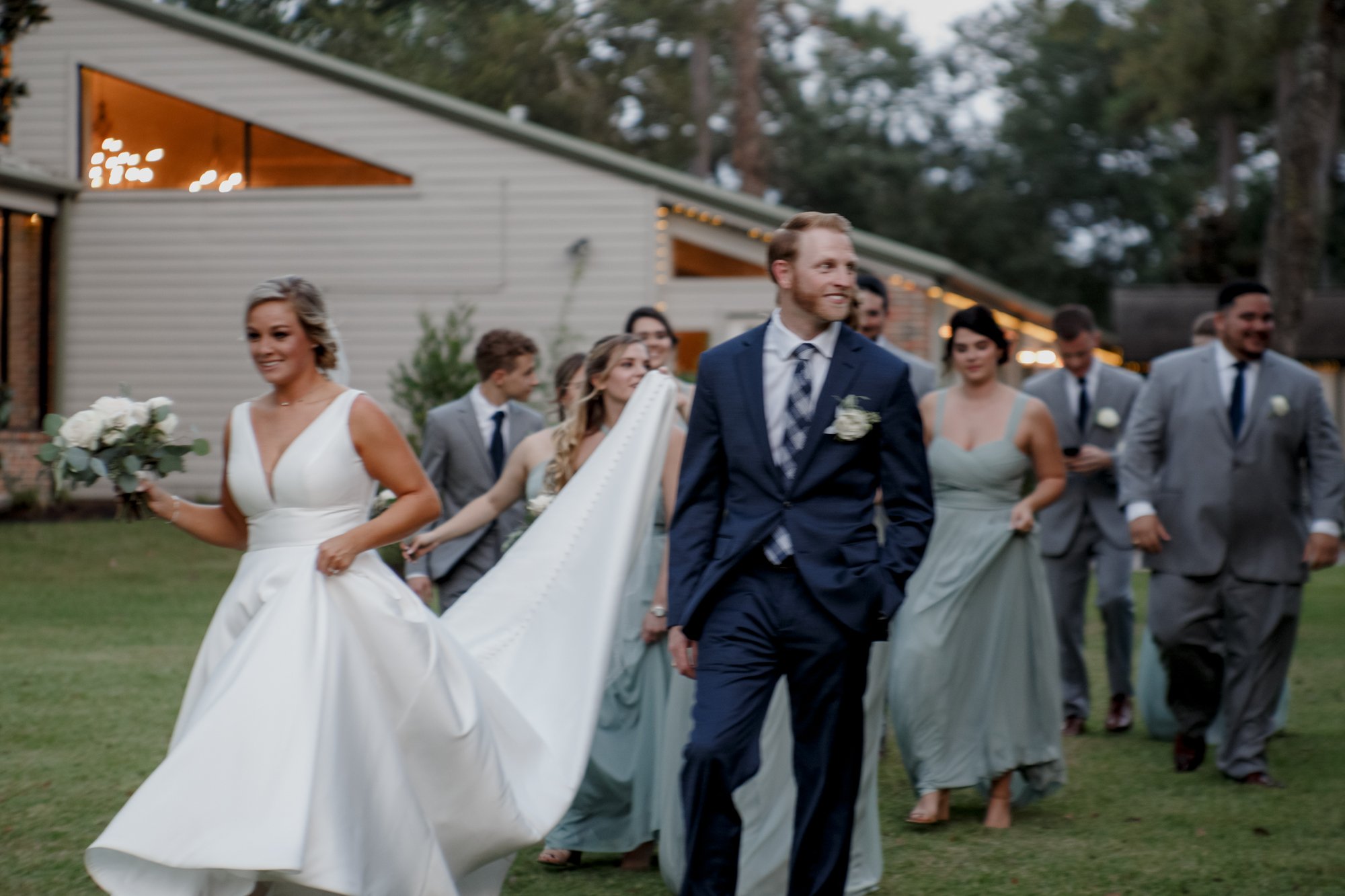 Groom carries bride's dress train with bridal party on the background. Wedding at Shirley Acres