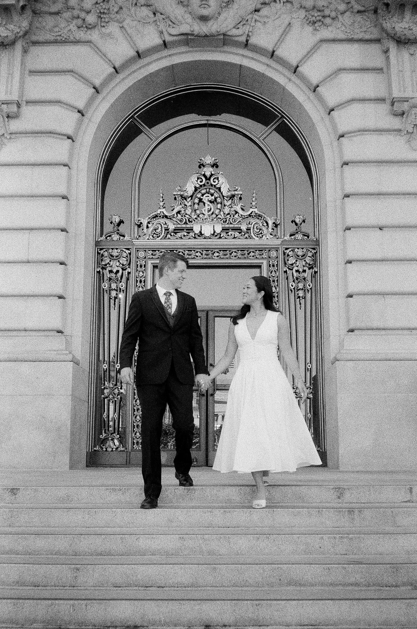 A man and a woman in wedding attire holding hands and walking down the stairs in front of ornate metal gates and a stone building with detailed architectural features.