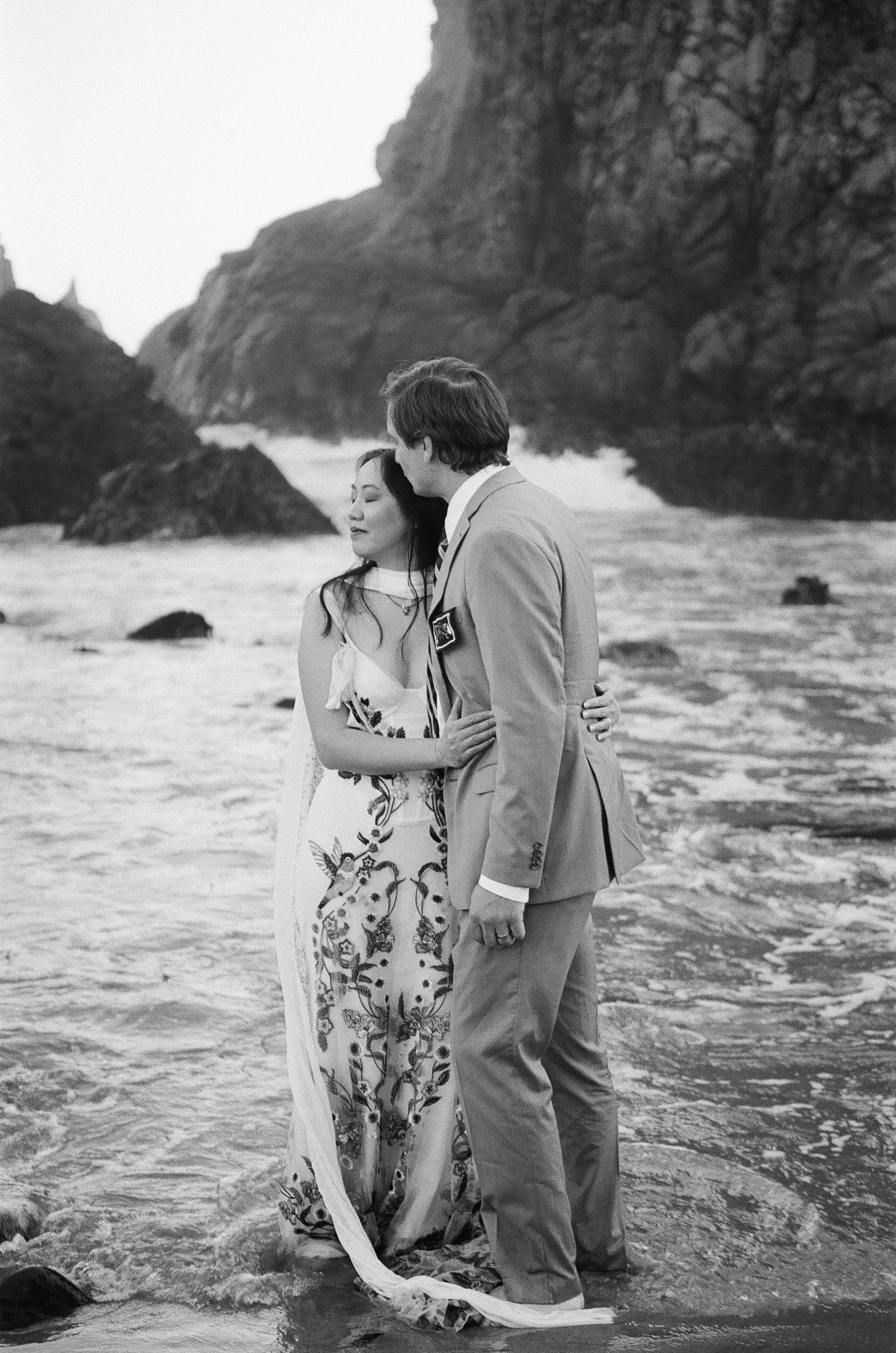 A black and white photo of a man and a woman standing close together on a beach, with rocky cliffs in the background. The woman is wearing a floral dress with her eyes closed, while the man is in a suit, gently kissing her on the head. They are embra