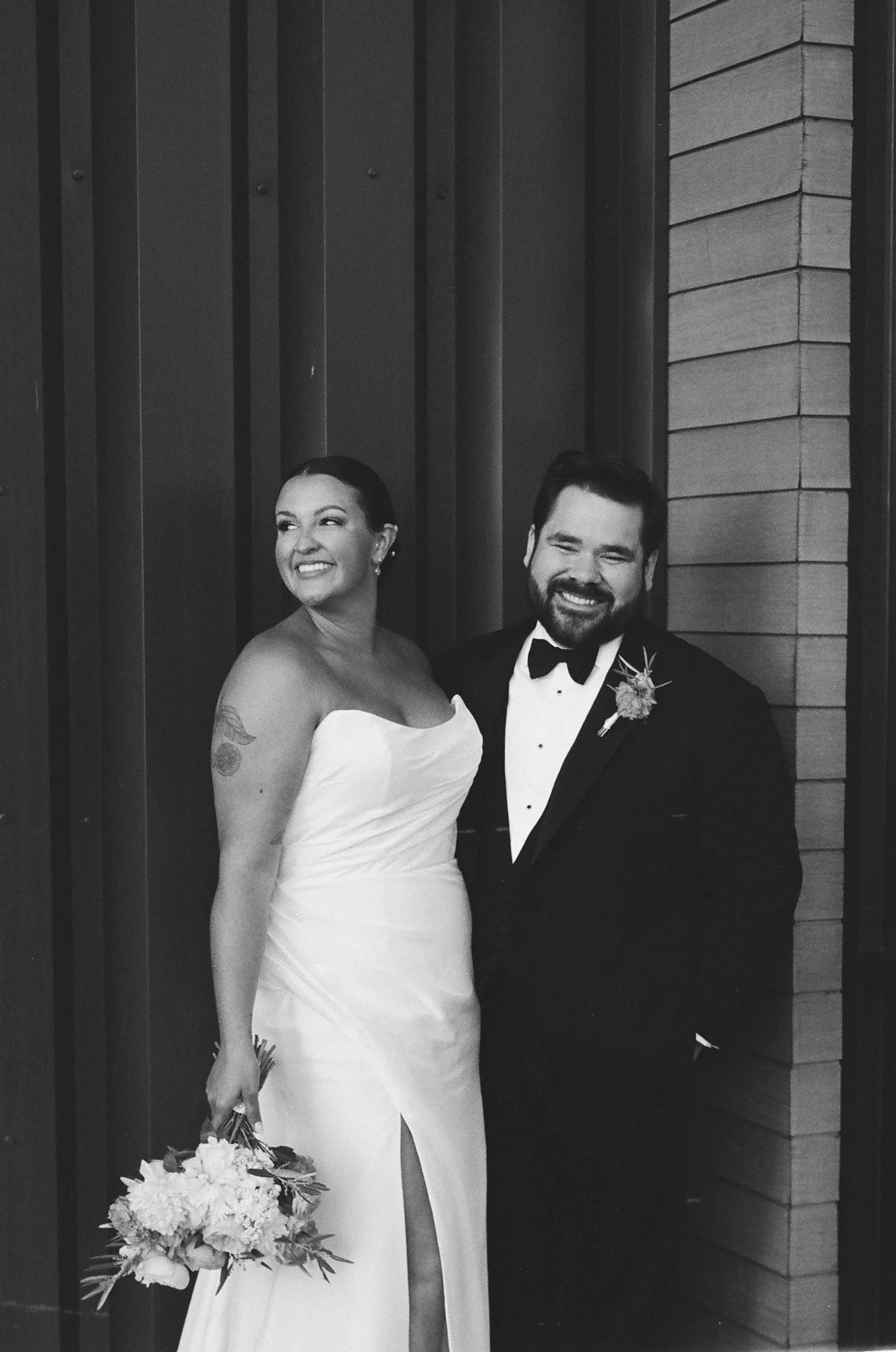 A black and white photo of a smiling bride and groom in wedding attire standing together in front of a wooden wall.