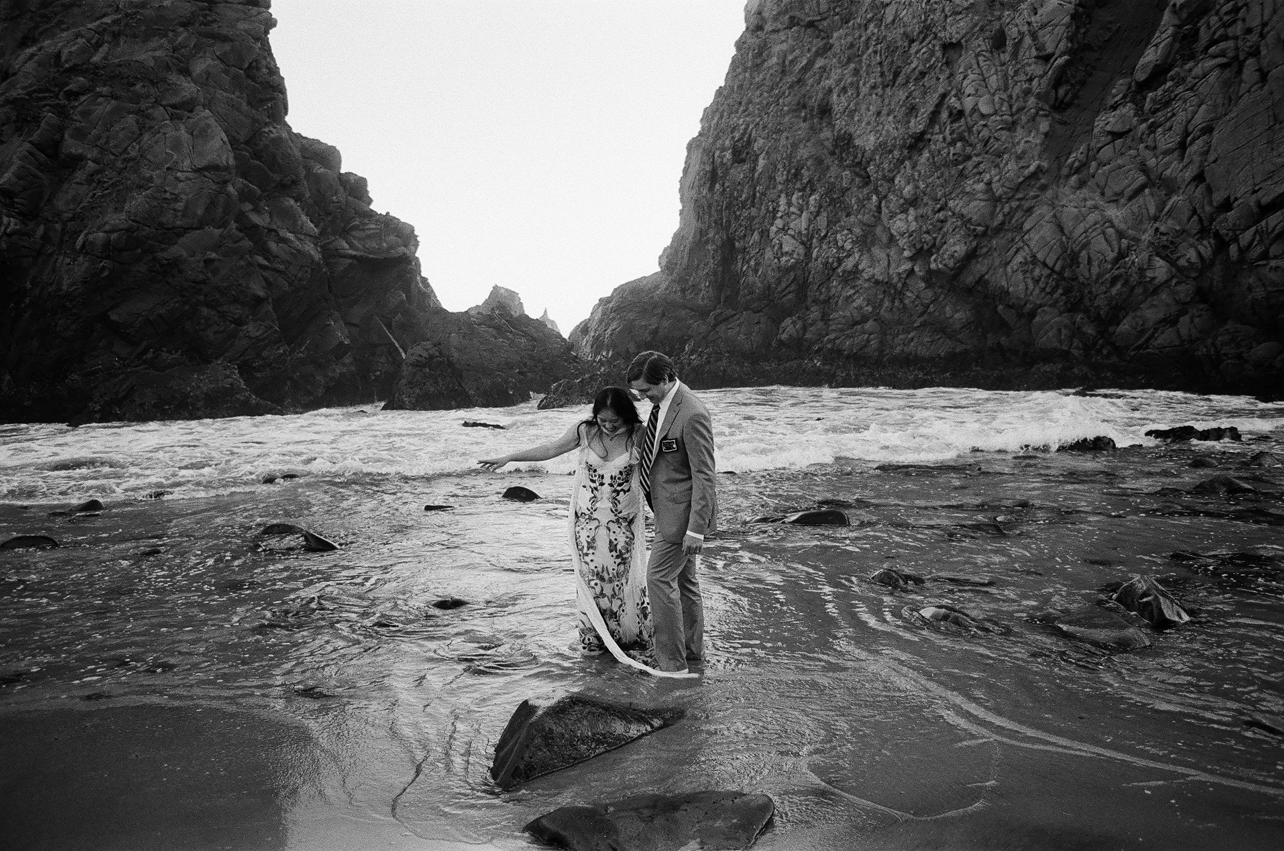 Black and white film photograph of couple embracing on rocky Big Sur Pfeiffer Beach between towering coastal cliffs. Bride wears embroidered floral gown as waves crash behind them. Moody California elopement by photographer Vivian Chen.