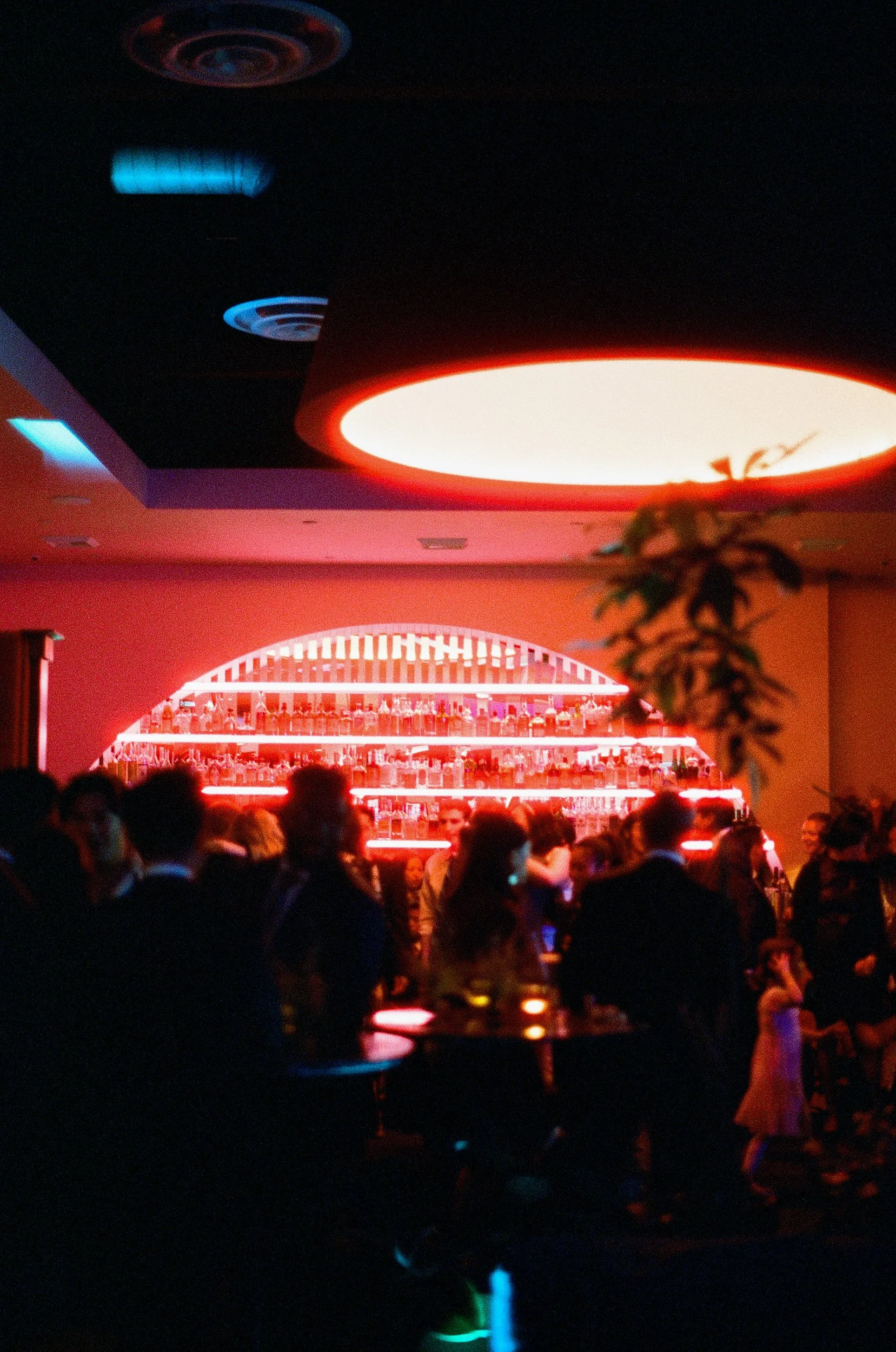 A crowded indoor bar with red and pink neon lighting, a large illuminated bar counter with bottles, and people socializing at the Moongate Lounge in Chinatown.