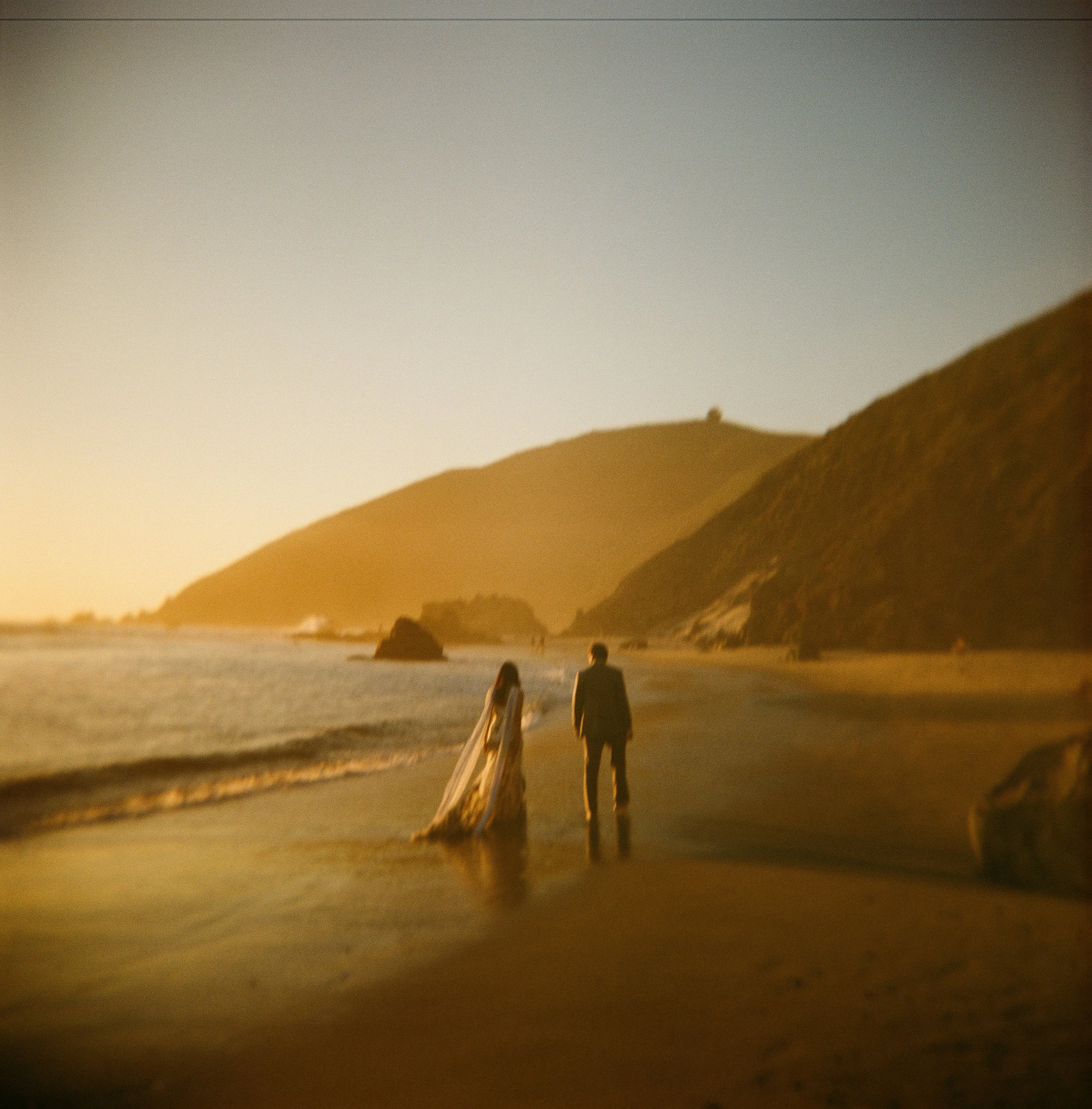 Newlyweds stroll along secluded Big Sur beach at sunset in this dreamy film photograph. Bride's flowing veil catches golden light as couple walks toward dramatic coastal cliffs. Intimate elopement photography by Vivian Chen.