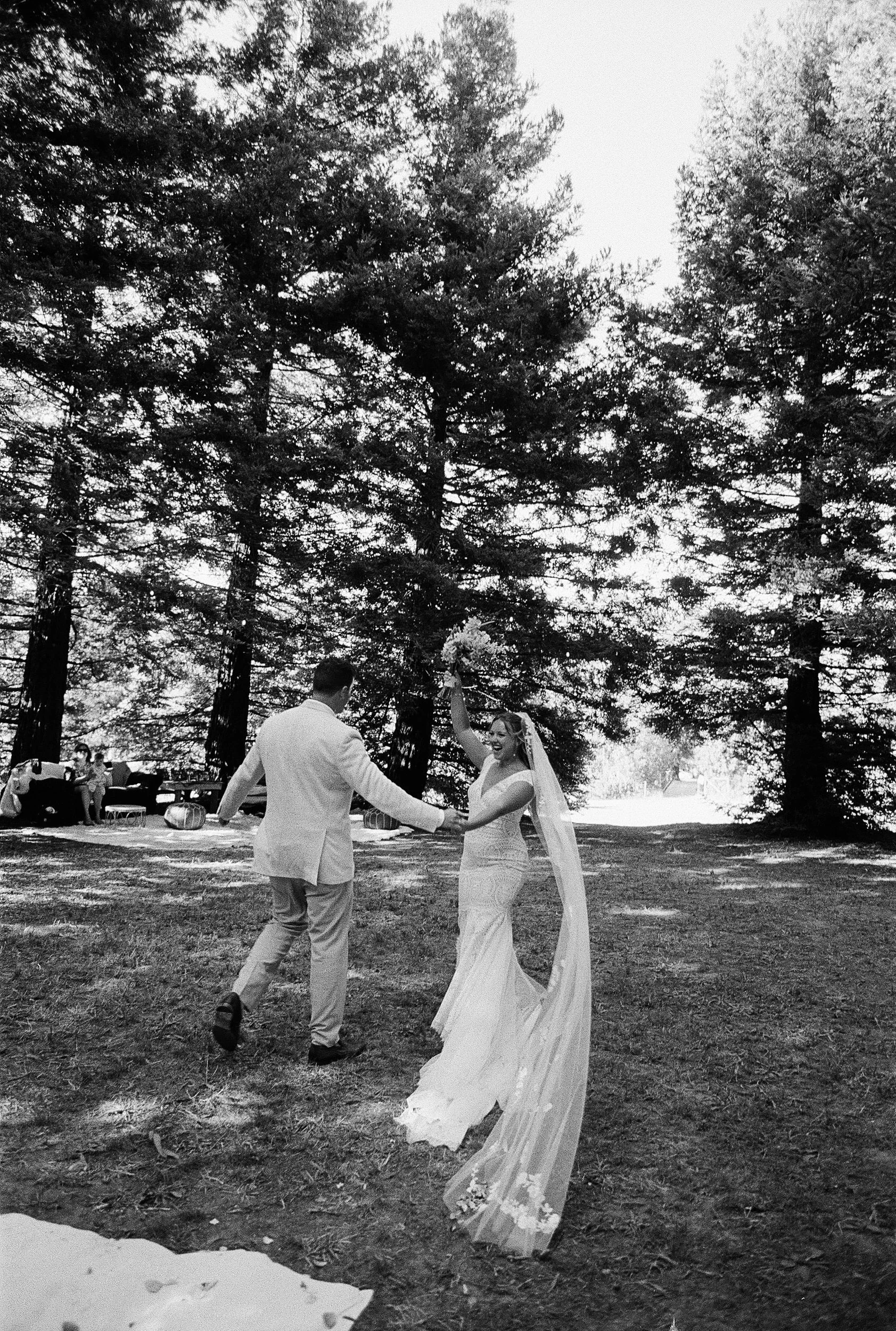 A bride and groom dancing outdoors under tall trees at a wedding reception, with guests seated in the background.