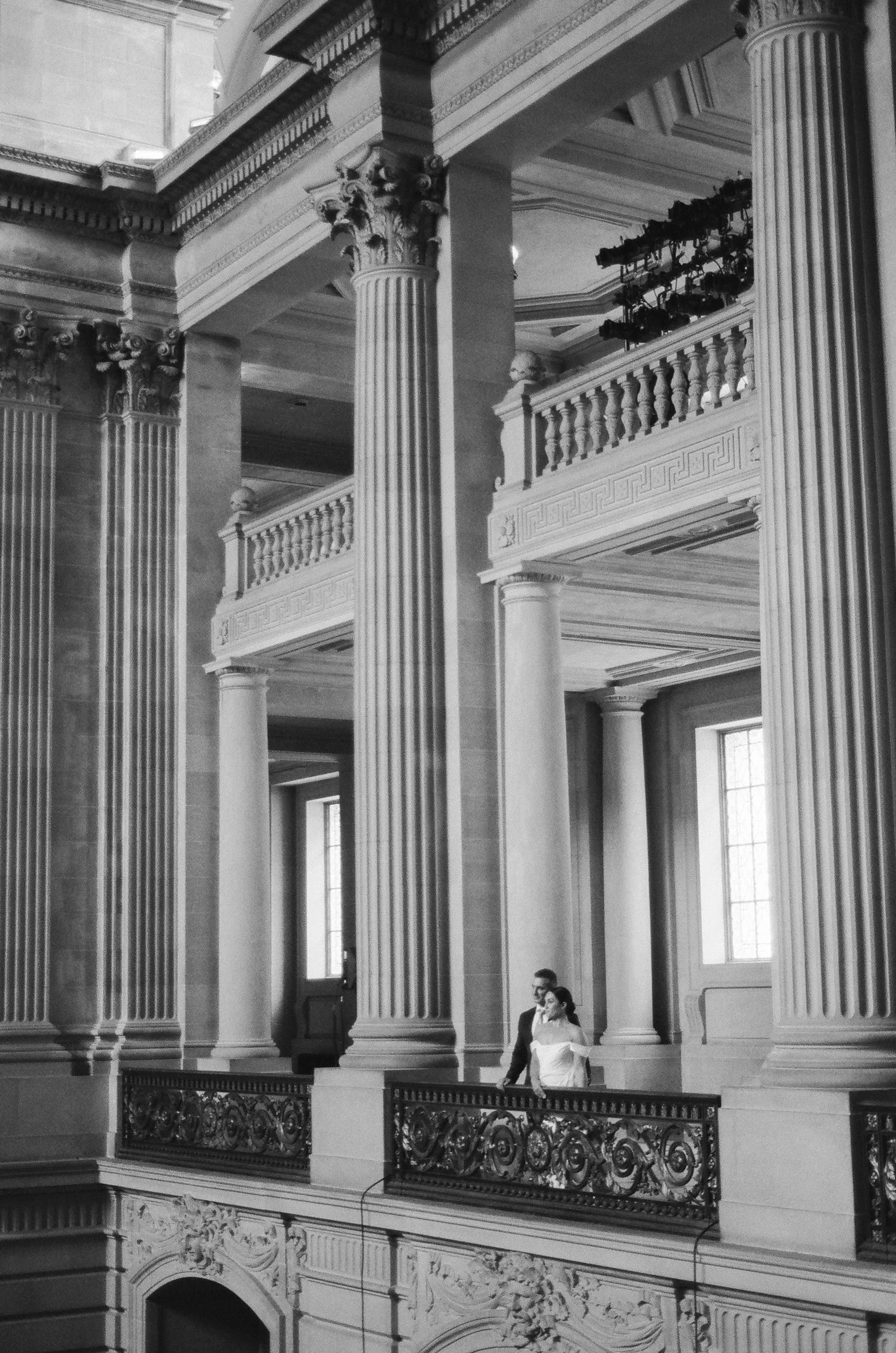 Interior of a grand building with tall Corinthian columns, ornate balustrades, and large windows. Two people are standing on a balcony, one man wearing a dark suit and woman in a light dress.