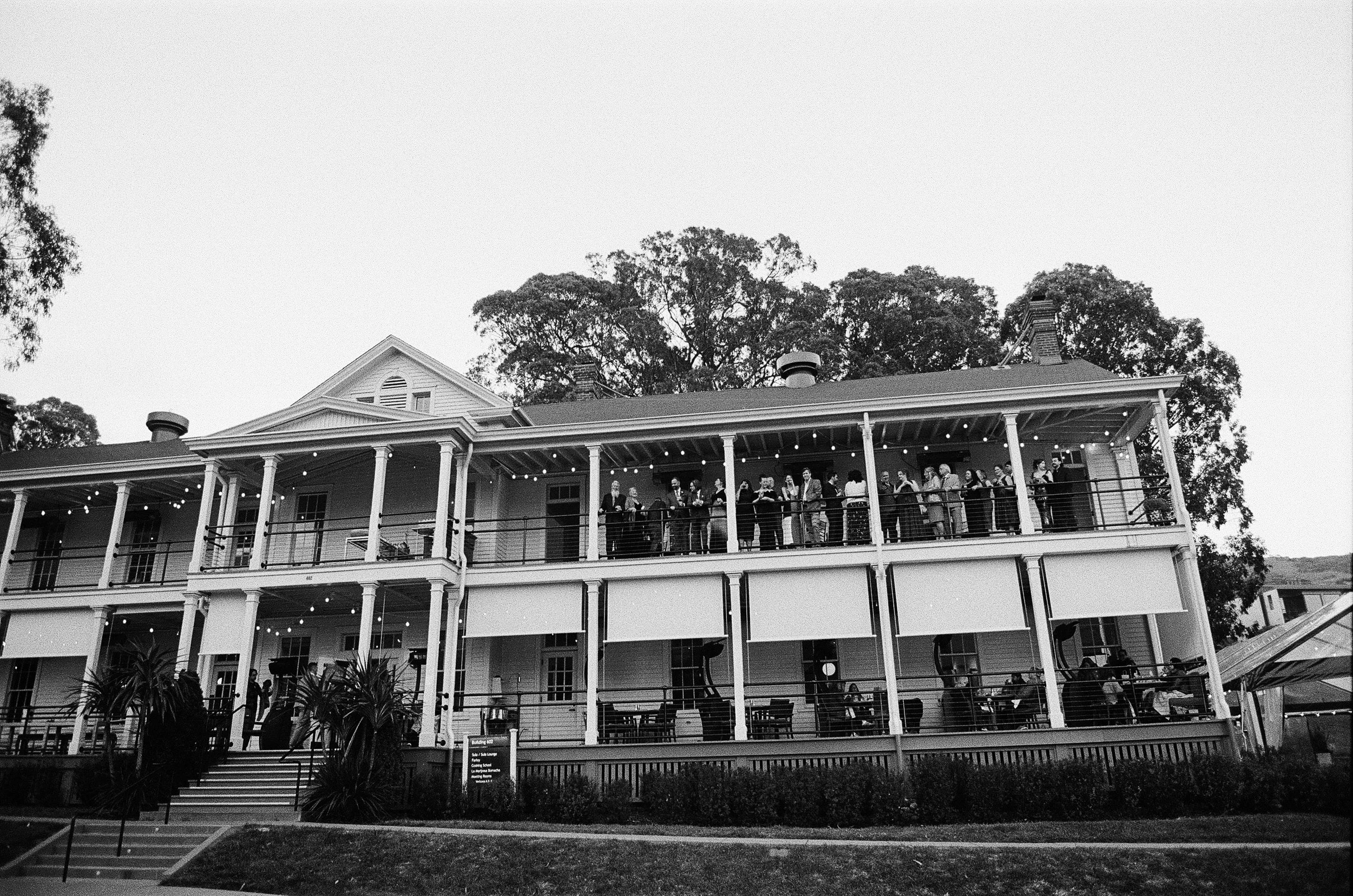 People gather on the balcony of a large, multi-story house with string lights hanging, trees in the background, and a sign in front of the stairs.
