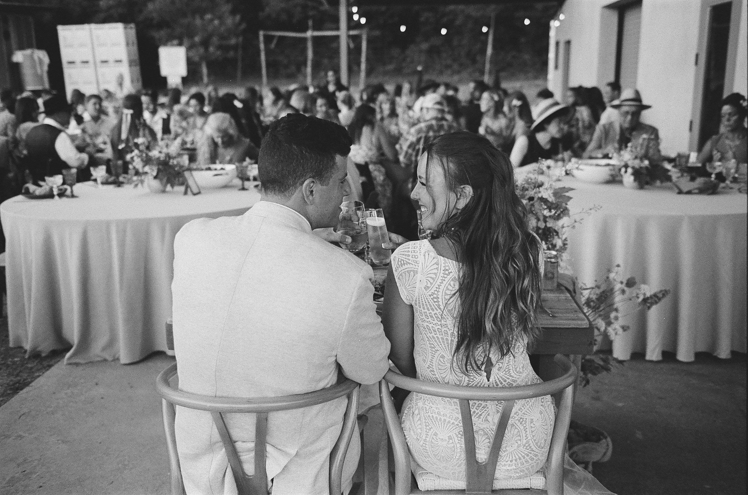 A black and white photo of a couple sitting closely and smiling at each other at a dinner party, with many guests seated at a long decorated table in the background.