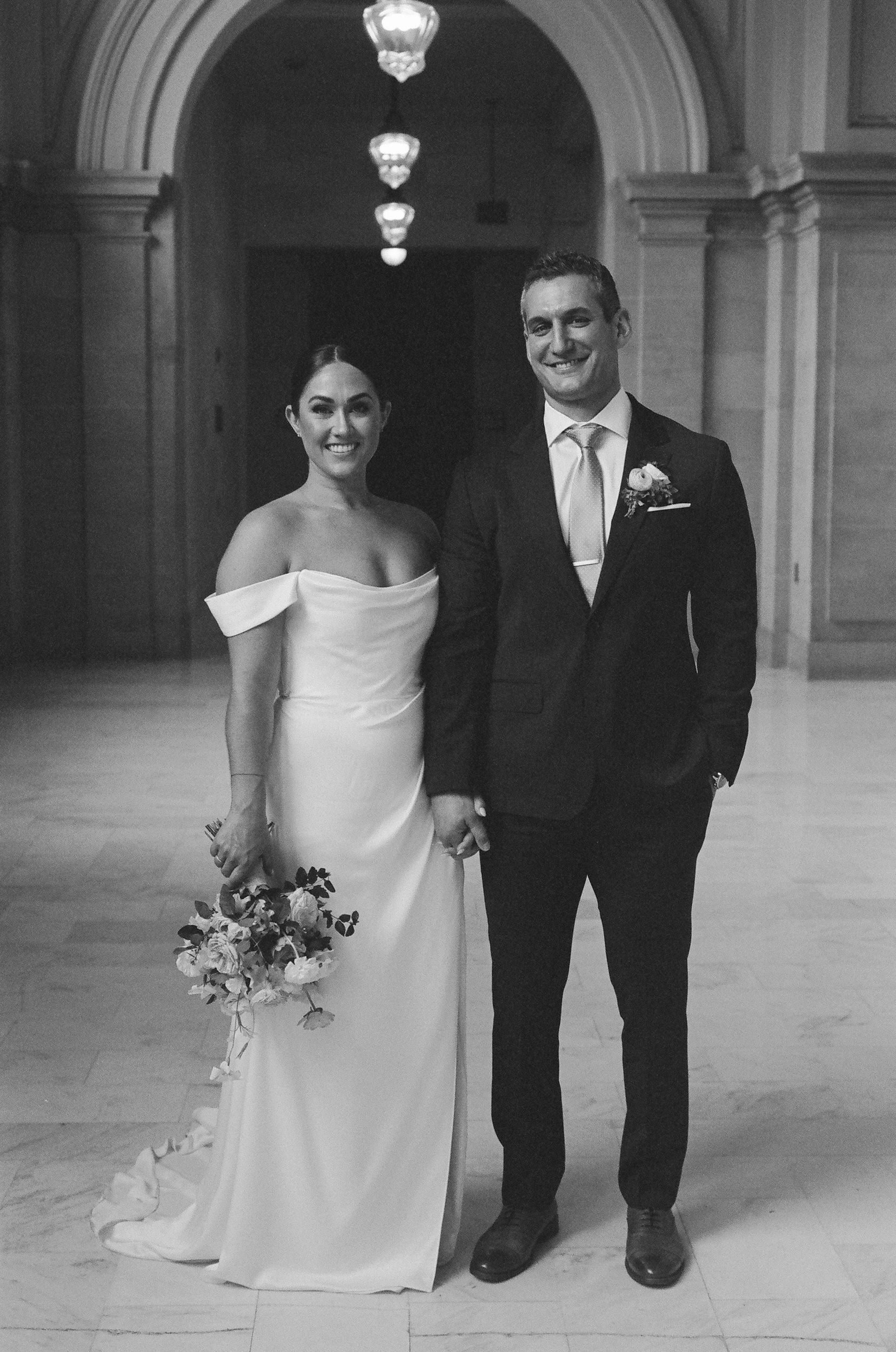 A black and white photo of a bride and groom standing together indoors. The bride is wearing a strapless wedding gown and holding a bouquet of flowers, while the groom is dressed in a suit with a boutonniere. They are smiling and holding hands.