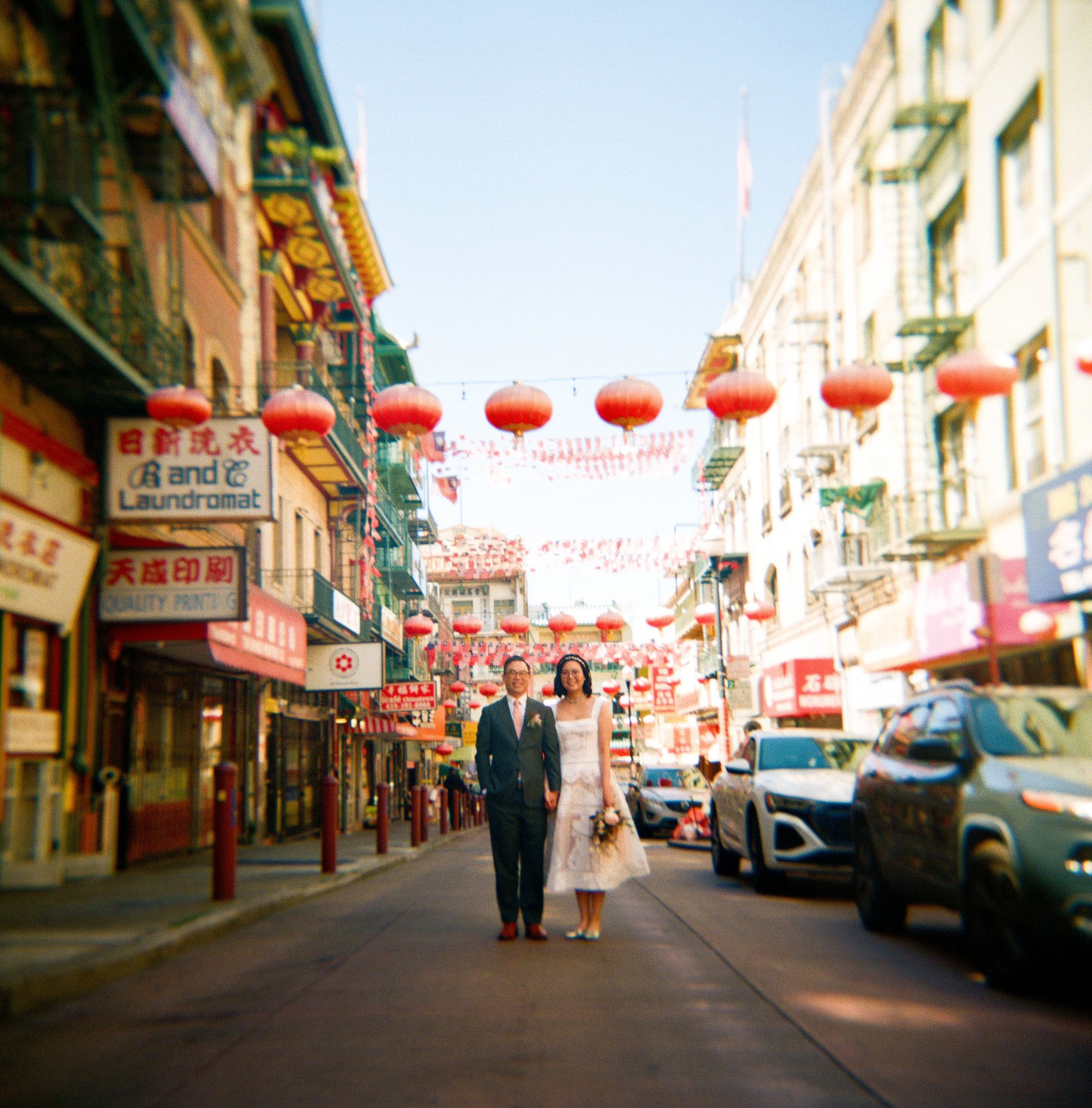 A man and woman walking down a busy Chinatown street adorned with red lanterns and colorful signage, with buildings on either side and cars parked along the street.