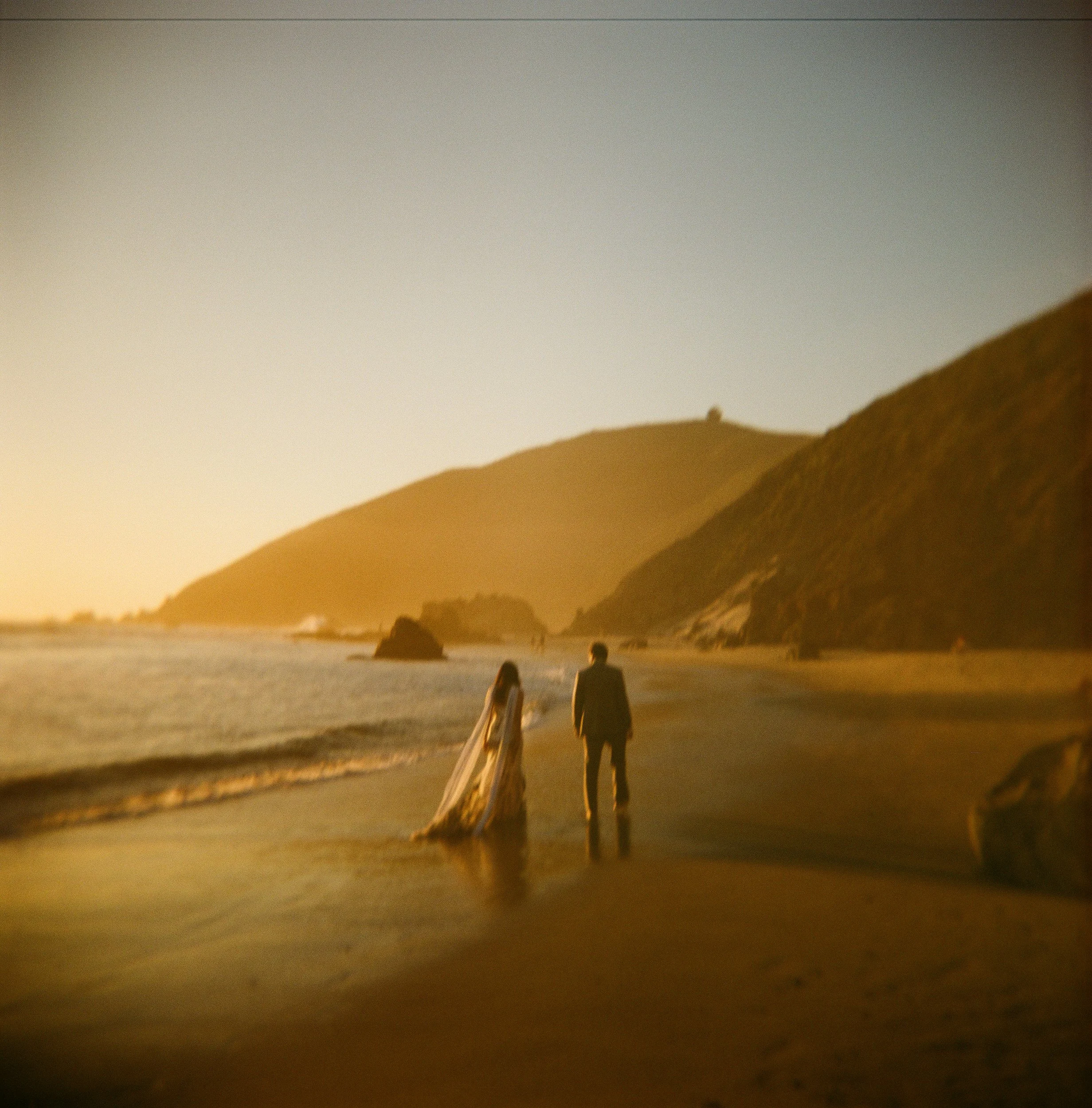 A landscape scene at sunset with silhouettes of two people walking along a sandy beach, grassy dunes in the background, and the sky transitioning from light to dark.