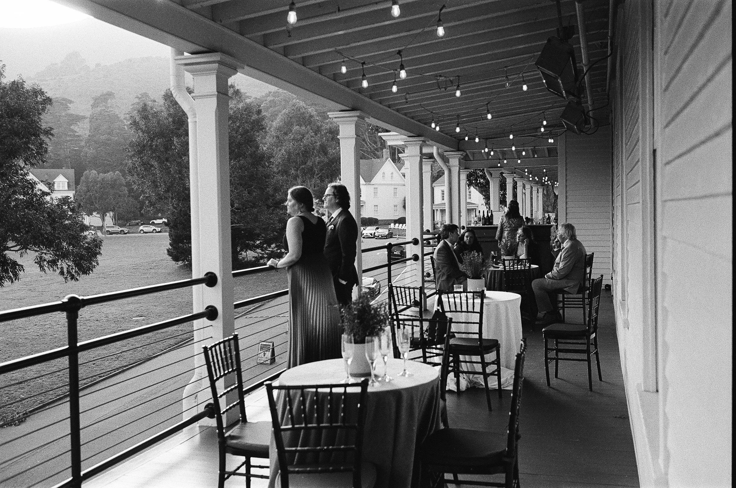 Black-and-white photo of an outdoor restaurant or cafe with tables and chairs, some with wine glasses and flower arrangements, overlooking a grassy area with trees and houses in the background. A couple stands on the porch, talking, as string lights 