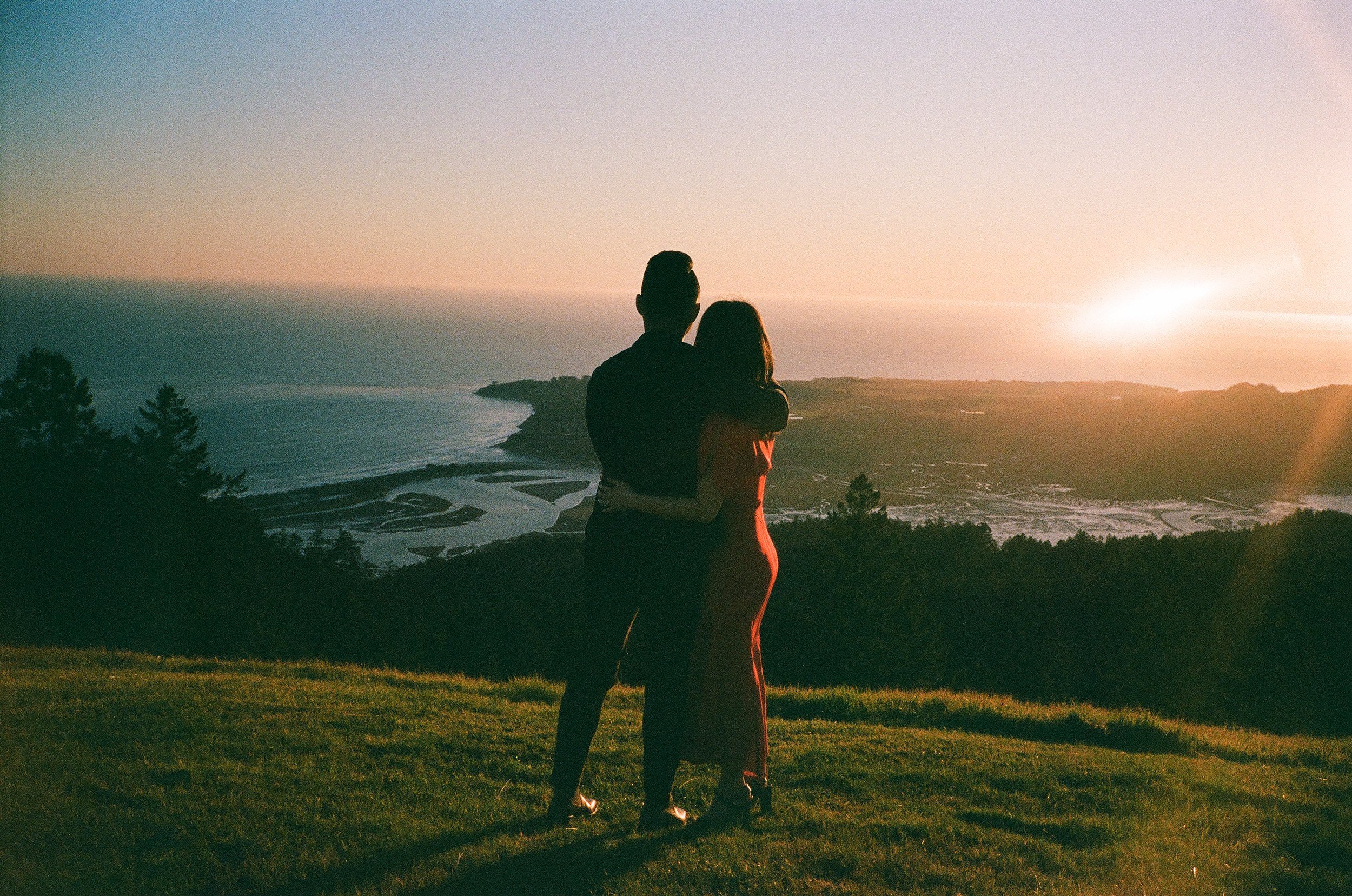 A silhouette of a couple embracing and watching a sunset over a scenic landscape with water, hills, and trees.