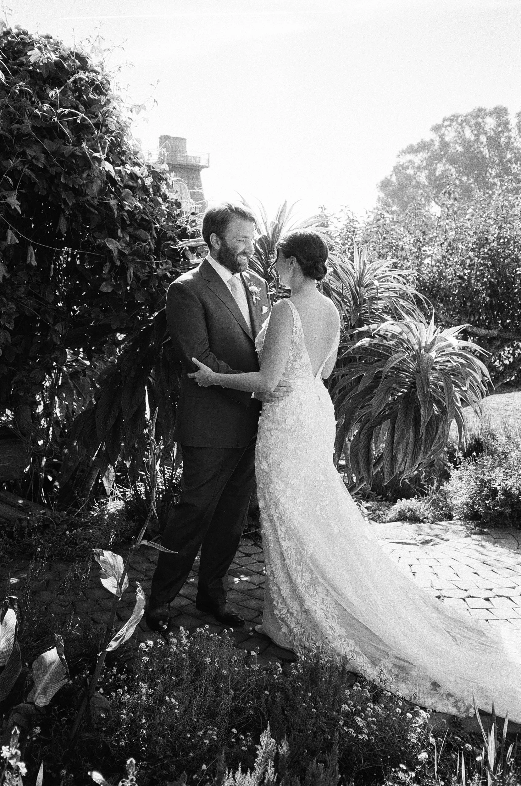 A black and white photo of a bride and groom holding hands and gazing at each other outdoors, surrounded by lush greenery.