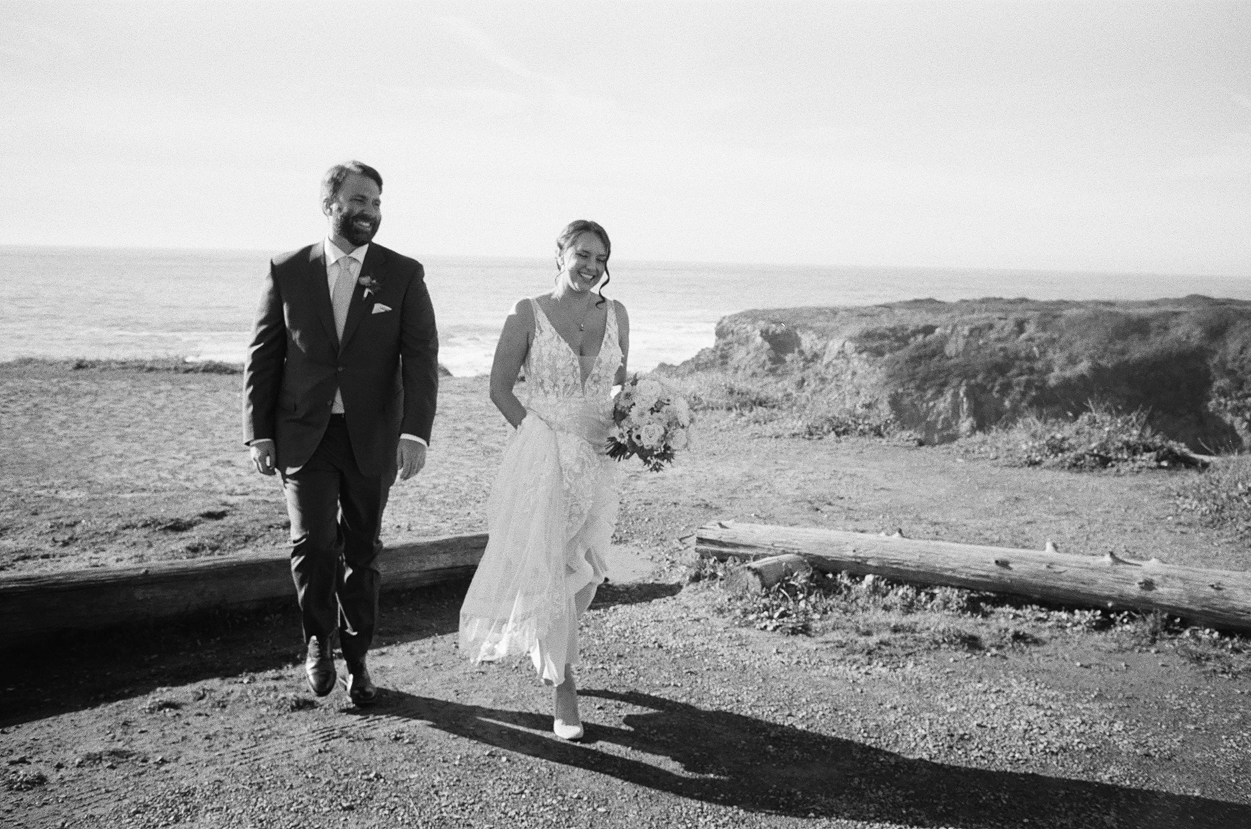 Black and white photo of a happy bride and groom walking along a beach with rocky cliffs in the background, the bride holding a bouquet of flowers.