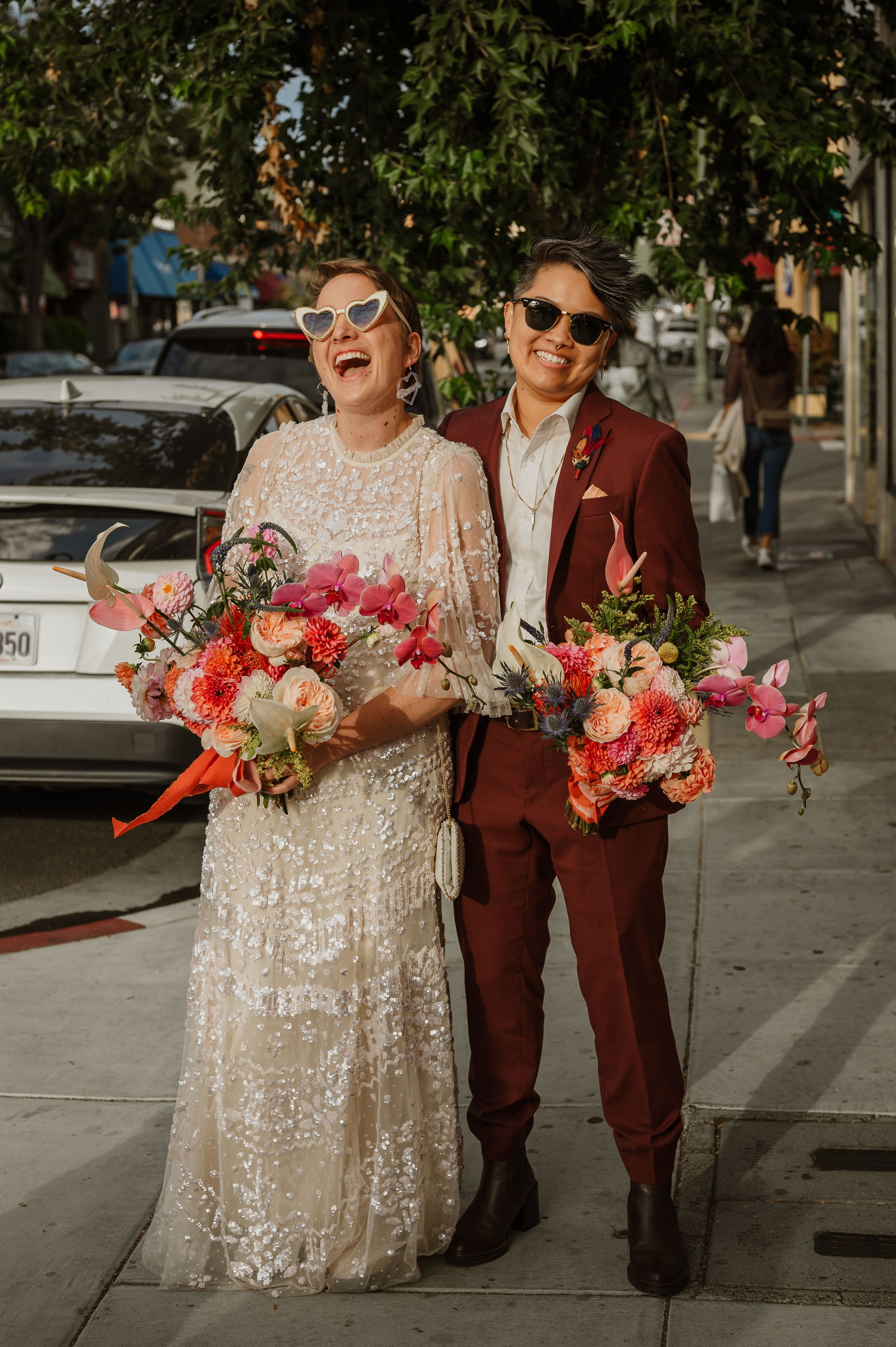 Two women standing on a city sidewalk, smiling and holding vibrant bouquets of flowers, dressed in formal attire with sunglasses, in daytime with a tree and parked cars in the background.