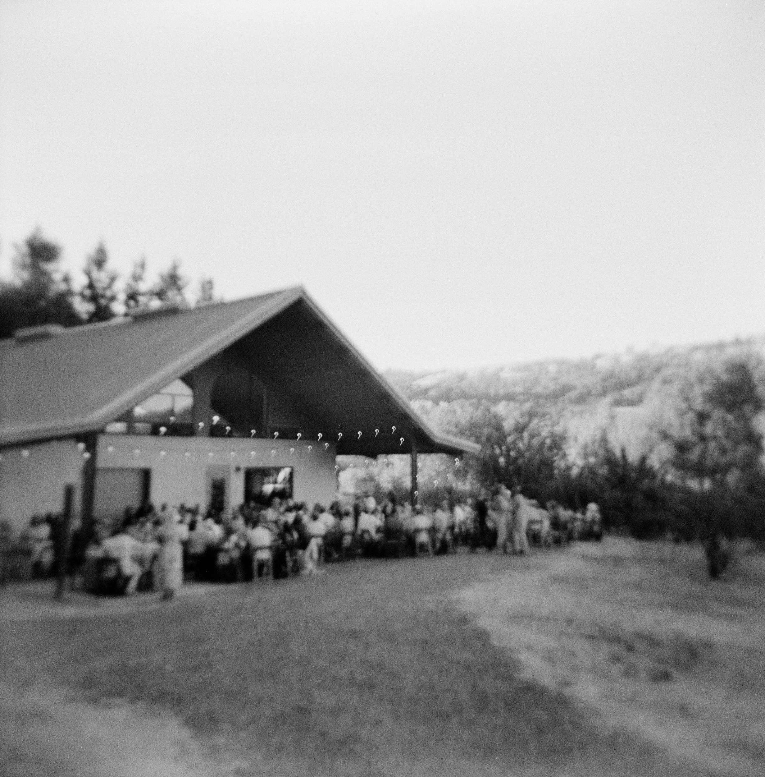 Black and white photo of a building with a triangular roof, trees, and a row of flowers in front.