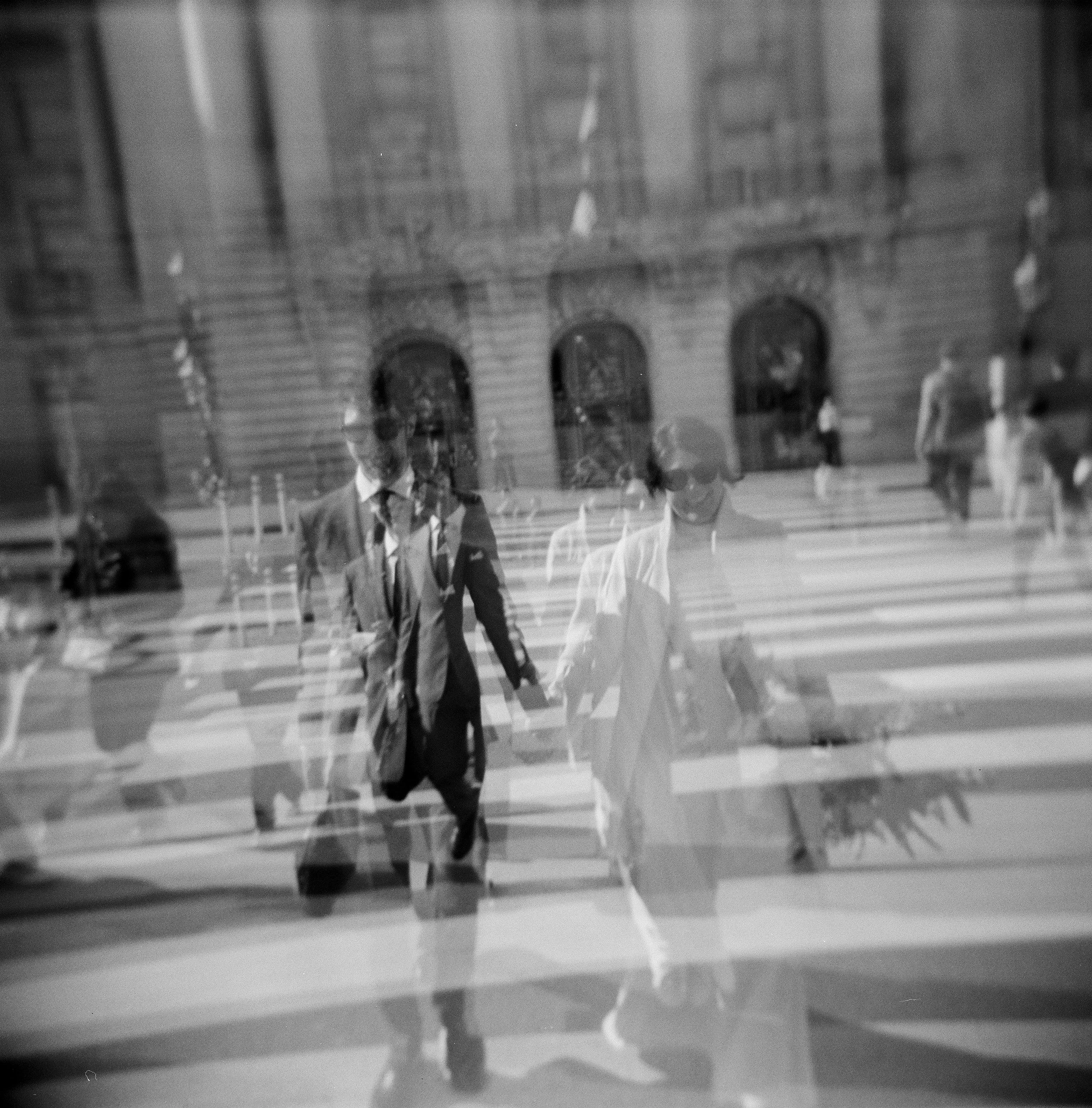Black and white double exposure Holga film photo of a San Francisco City Hall elopement couple holding hands crossing a city street with a reflection of the scene on the glass.