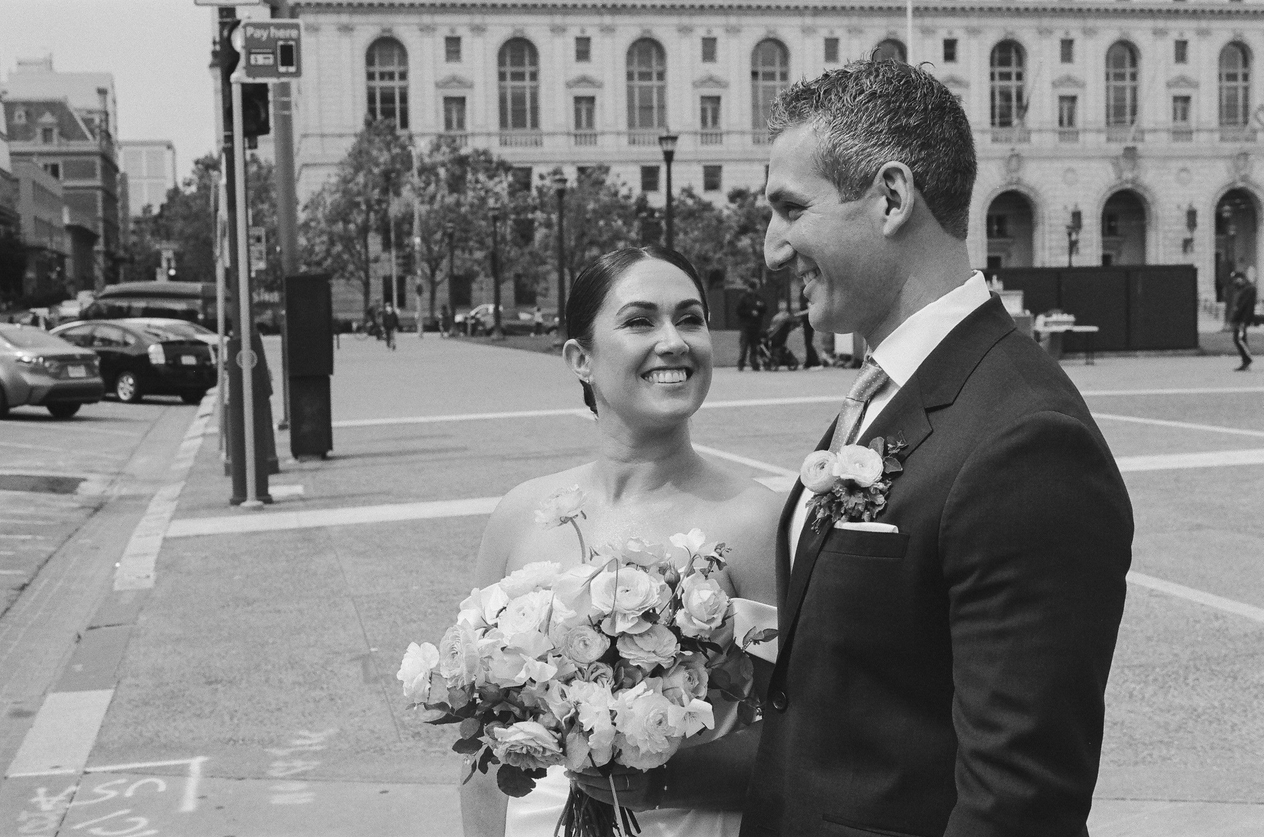 A smiling woman in a wedding dress holding a bouquet, standing close to a man in a tuxedo with a boutonniere, outside in an urban area with historical buildings and parked cars in the background.