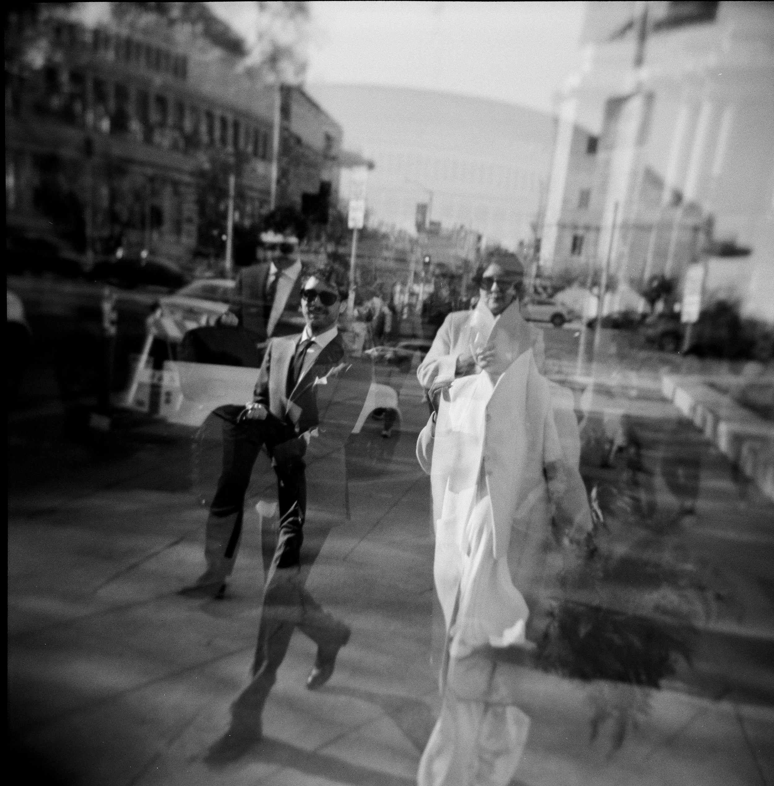 Black and white double exposure Holga film photo of a couple walking outside of SF City Hall, seen through a window with reflections of buildings, cars, and other objects.
