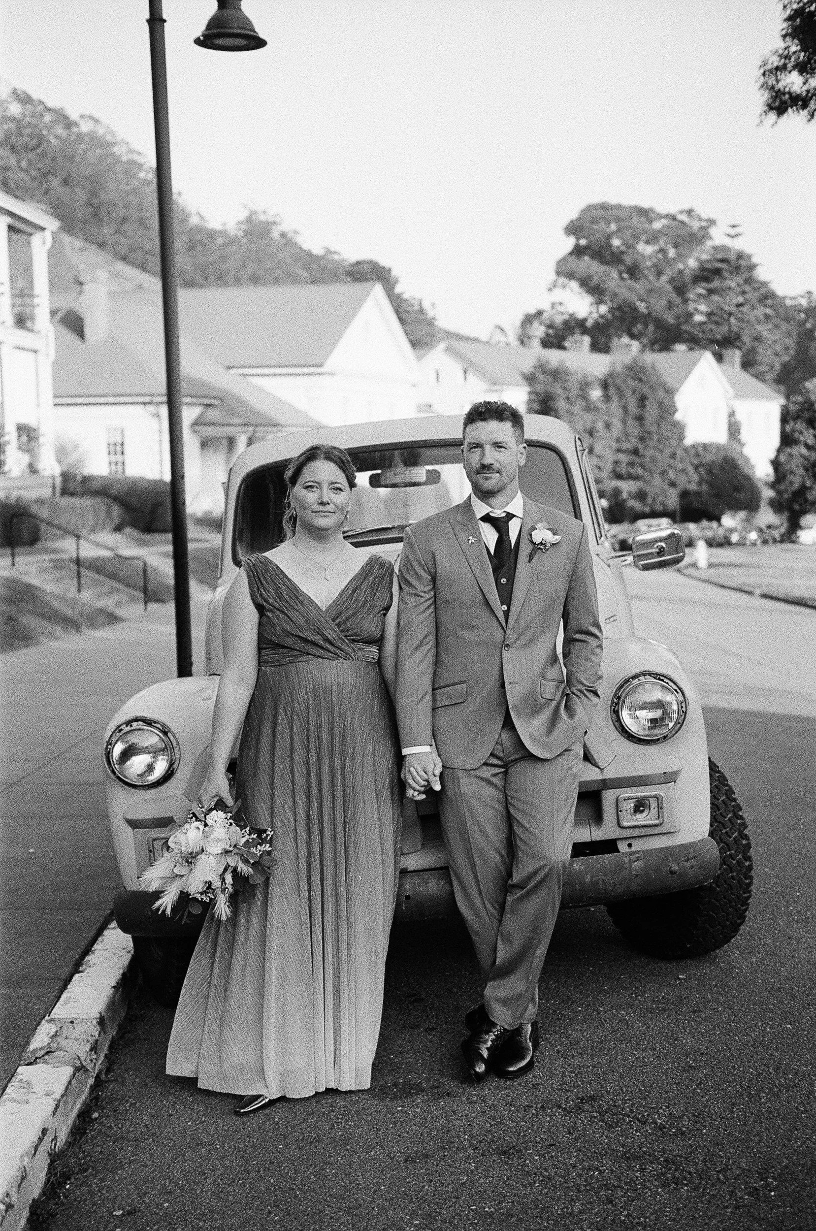 A black and white photo of a bride and groom standing in front of a vintage truck on a street. The bride holds a bouquet and wears a long gown; the groom is in a suit. They are holding hands and looking at the camera.