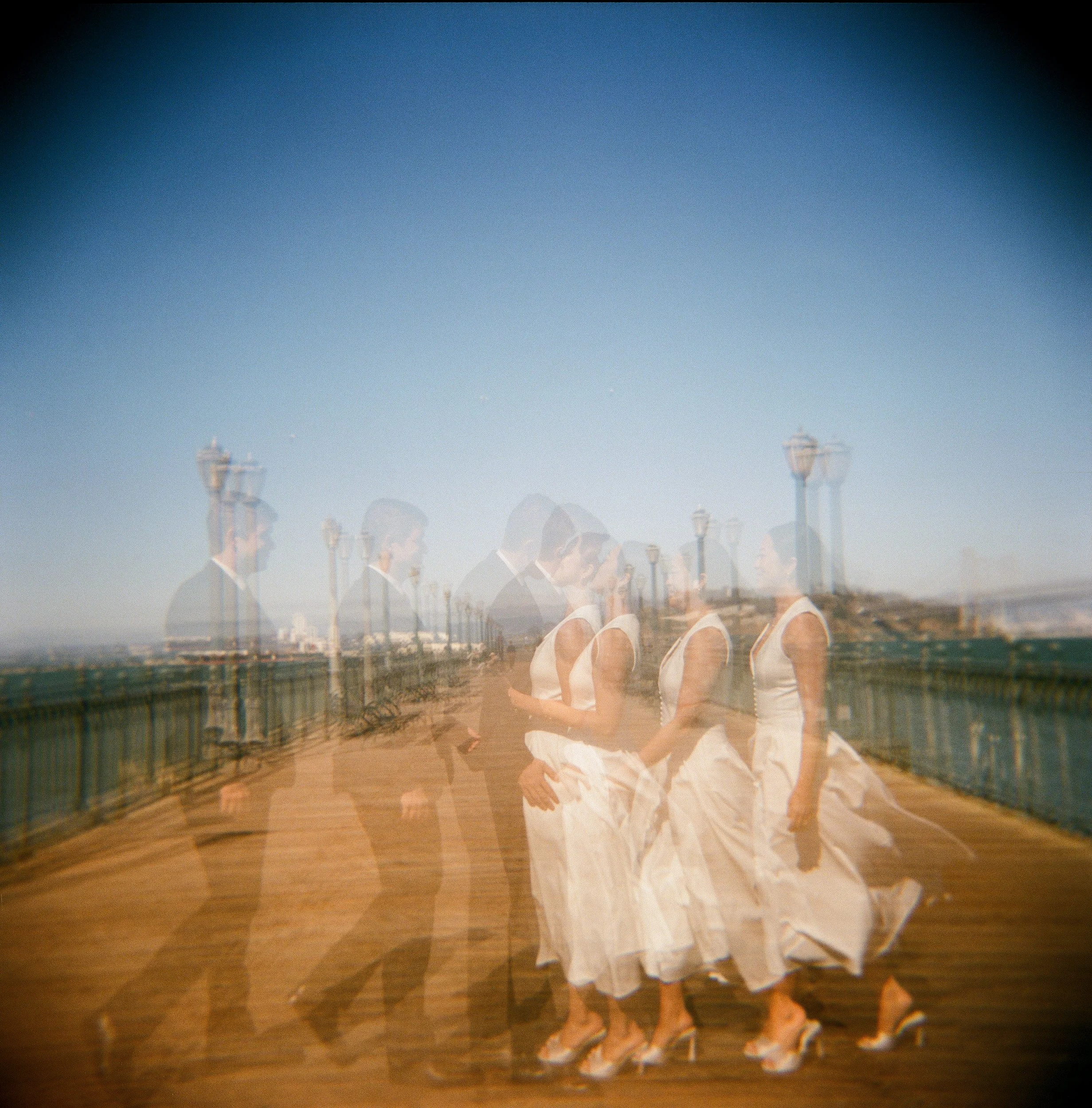 A double exposure photograph showing a person wearing a white dress with floral details, standing outdoors in daylight with a clear blue sky and some structures in the background.