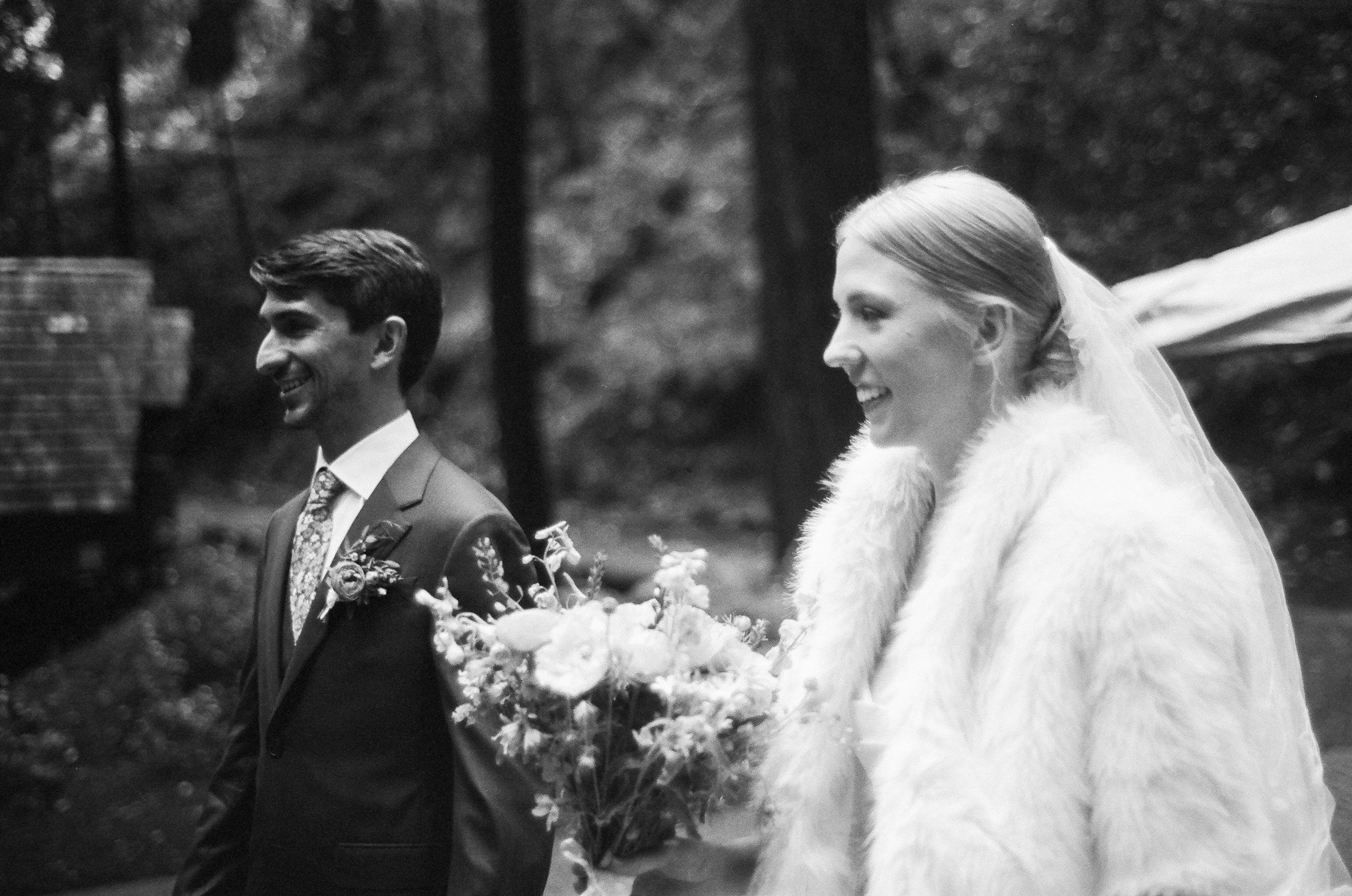 Black and white photo of a man and a woman at what appears to be a wedding or special occasion, walking outdoors among trees. The man is wearing a suit with a floral tie, and the woman is wearing a dress with a fur shawl and is holding a bouquet of f