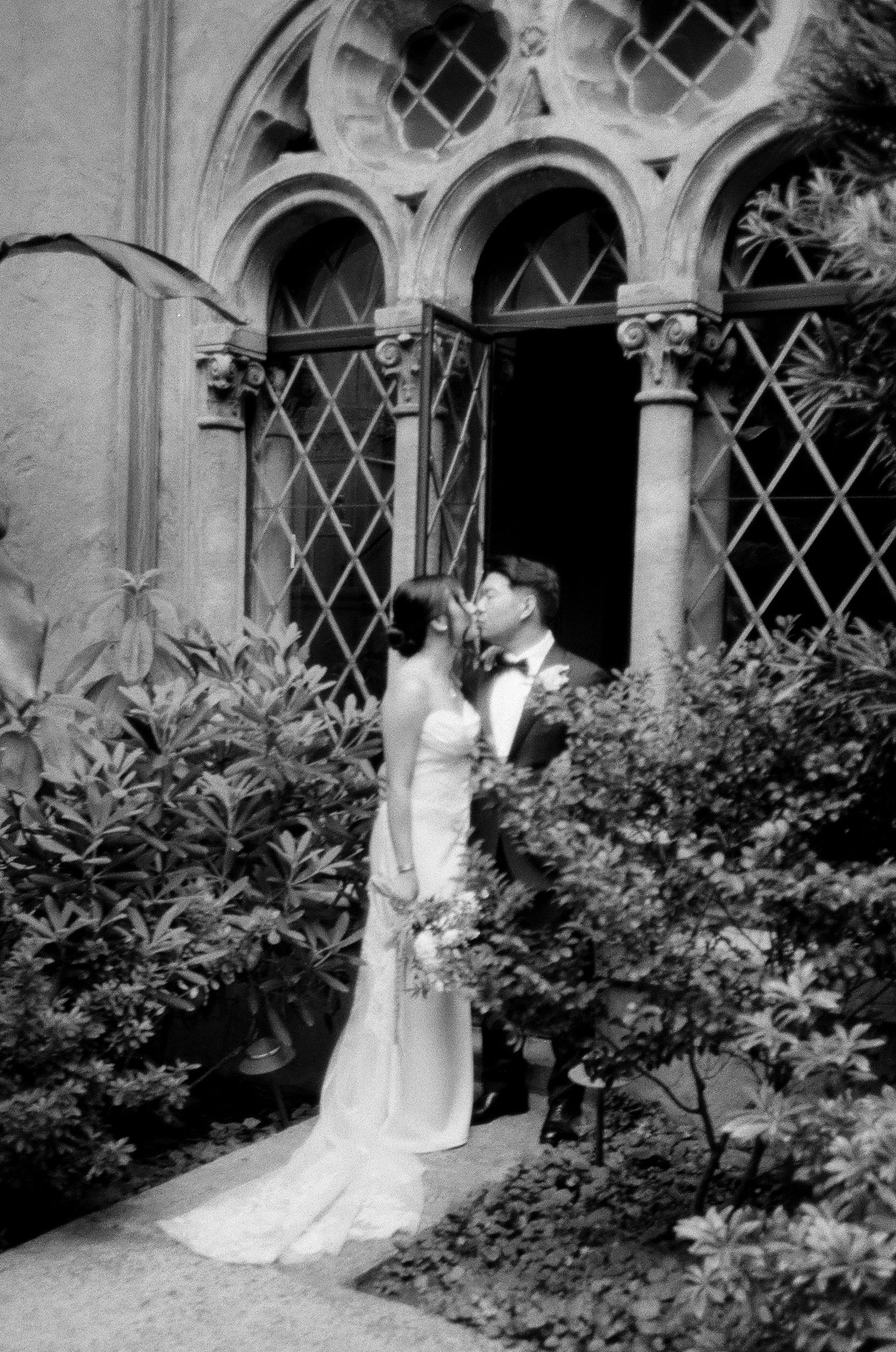 A black and white photo of a couple kissing in front of an ornate building with arched windows, surrounded by lush bushes and plants, with a pathway underneath. Photographed on black and white film using a Contax T2.