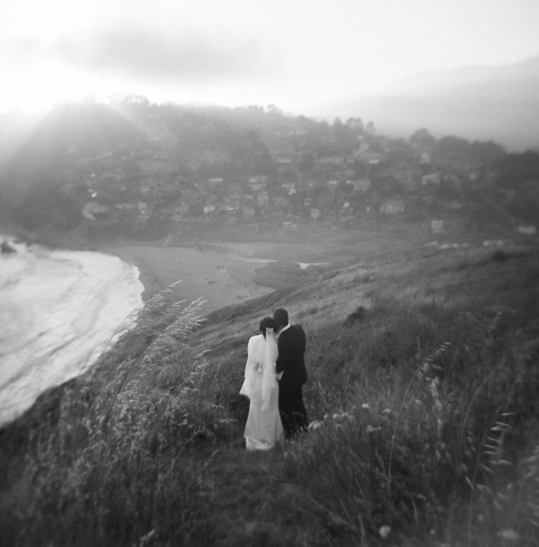 A black and white photo of a large wave crashing onto the beach with two people standing on the grass, facing the ocean.