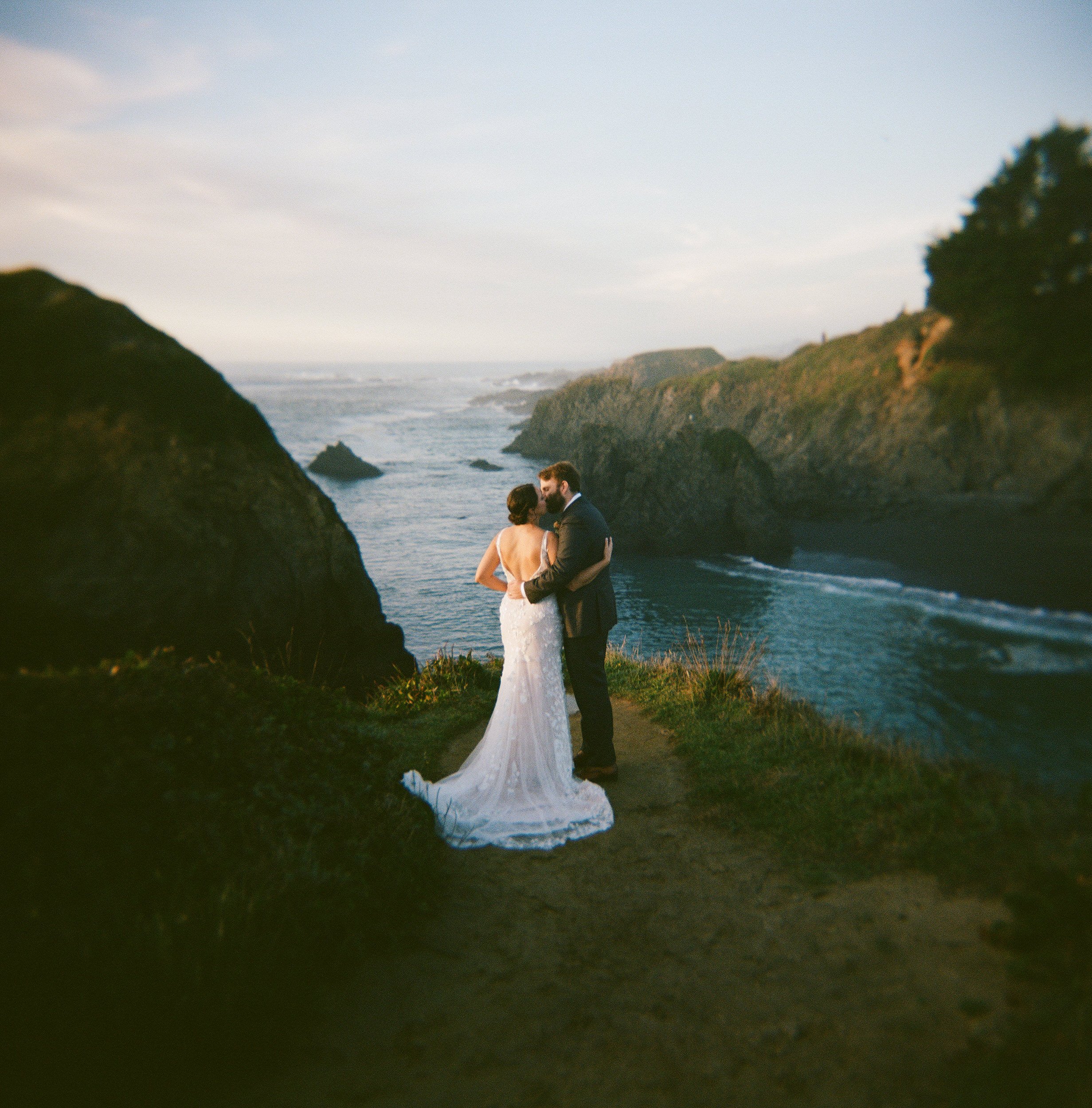 A couple dressed in wedding attire, standing on a grassy area near the ocean, with cliffs and overcast sky in the background.