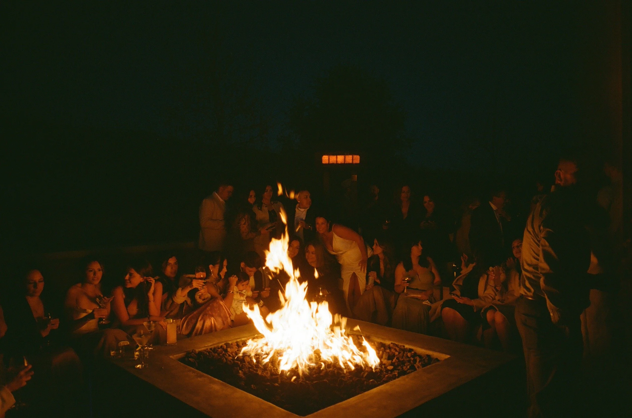 Group of people gathered around a fire pit at night, socializing and enjoying drinks at an outdoor event.